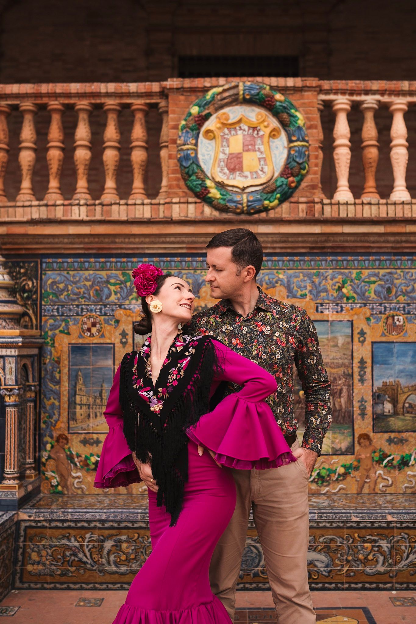 Couple embracing before colorful mosaic tiles at Plaza de España Seville, woman in flamenco dress and man in floral shirt.