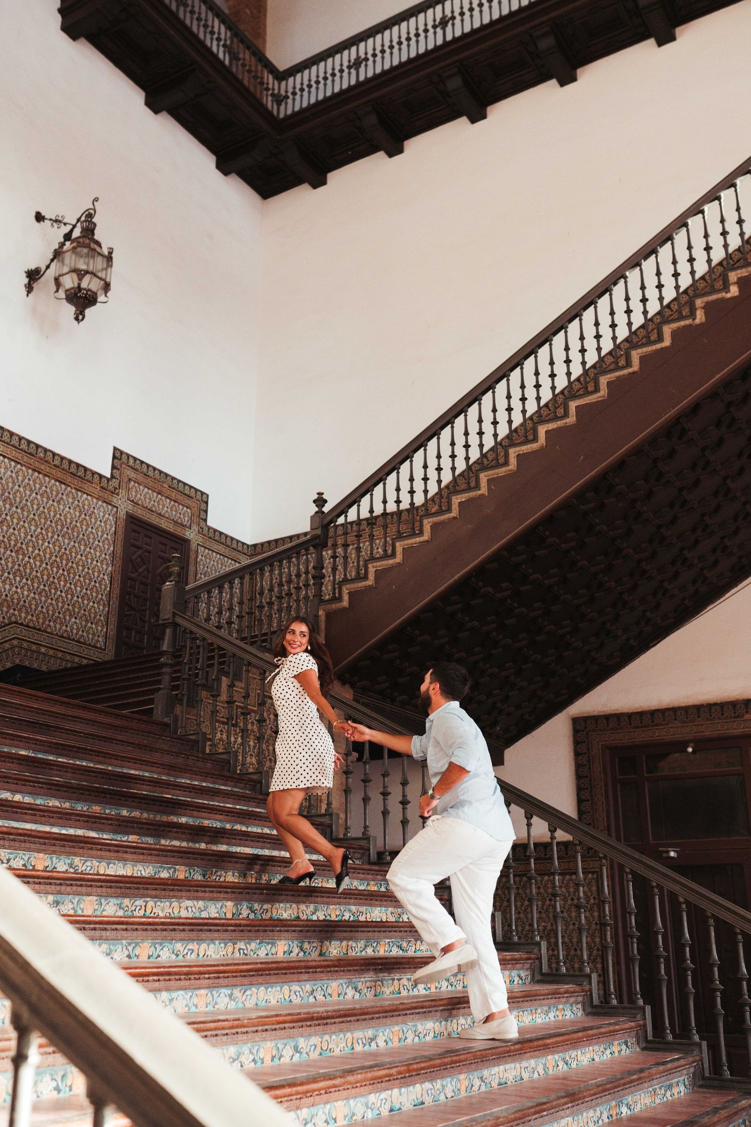 Couple holding hands on ornate tiled staircase with dark wood railings in elegant historic building.