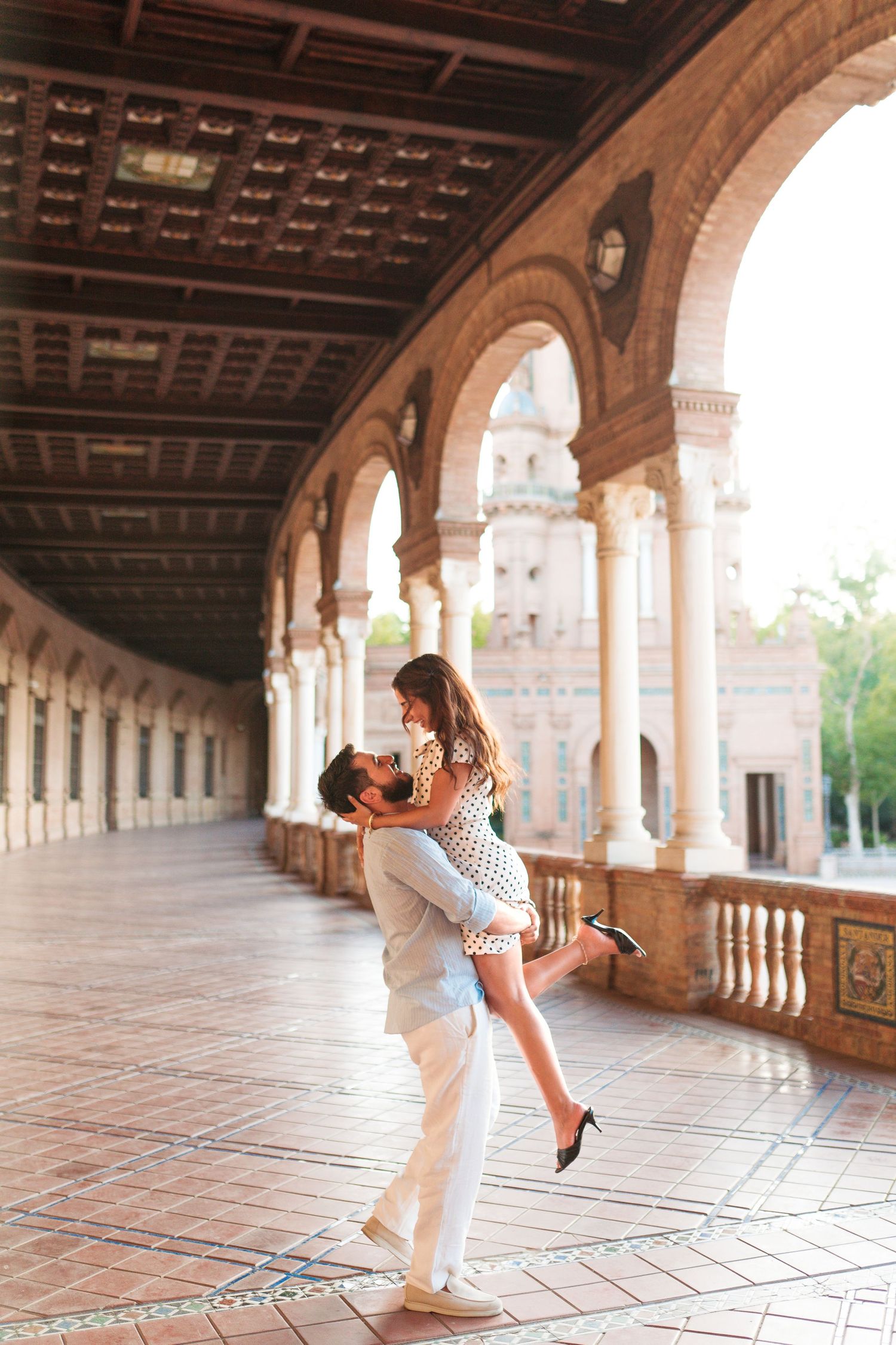 Romantic couple embracing in a sunlit Spanish plaza arcade with ornate arches and decorative ceiling.