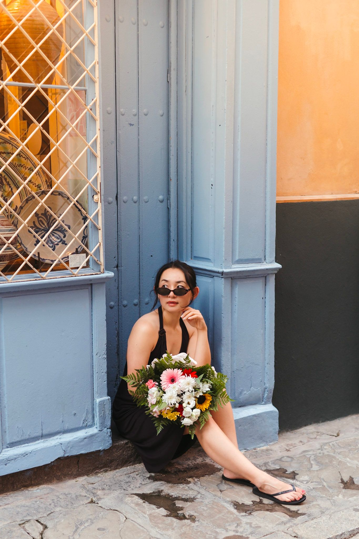 Stylish Asian woman in black dress holding flower bouquet sitting by blue door on cobblestone street.