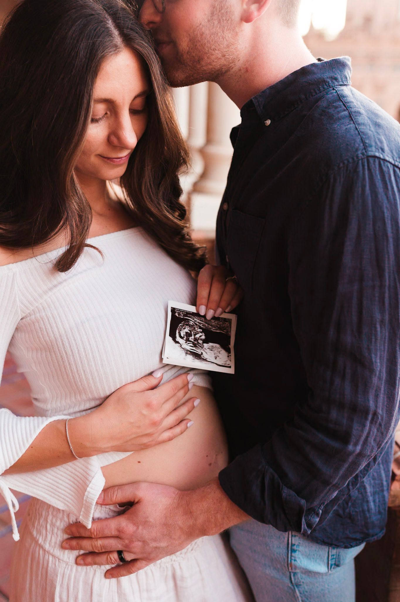 Couple embraces tenderly holding ultrasound photo against pregnant belly.