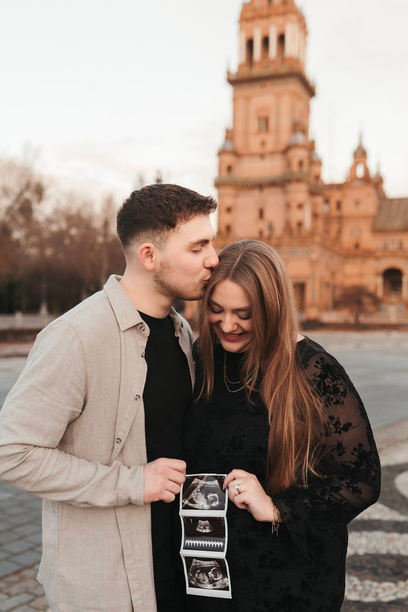 Man kisses pregnant woman's head as they hold ultrasound photos with historic cathedral behind them.