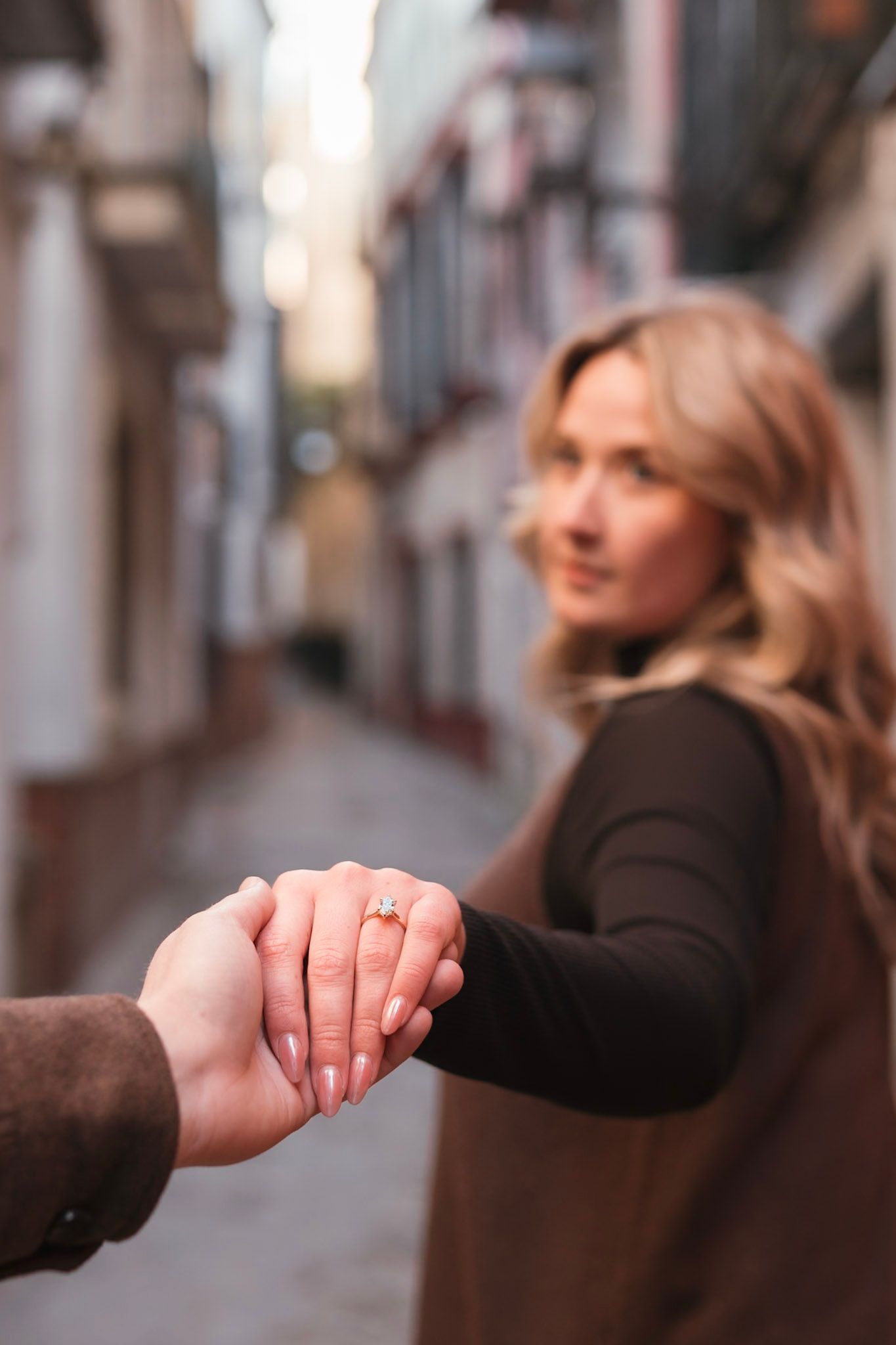 Newly engaged woman showing diamond ring while holding fiance's hand on charming European street.