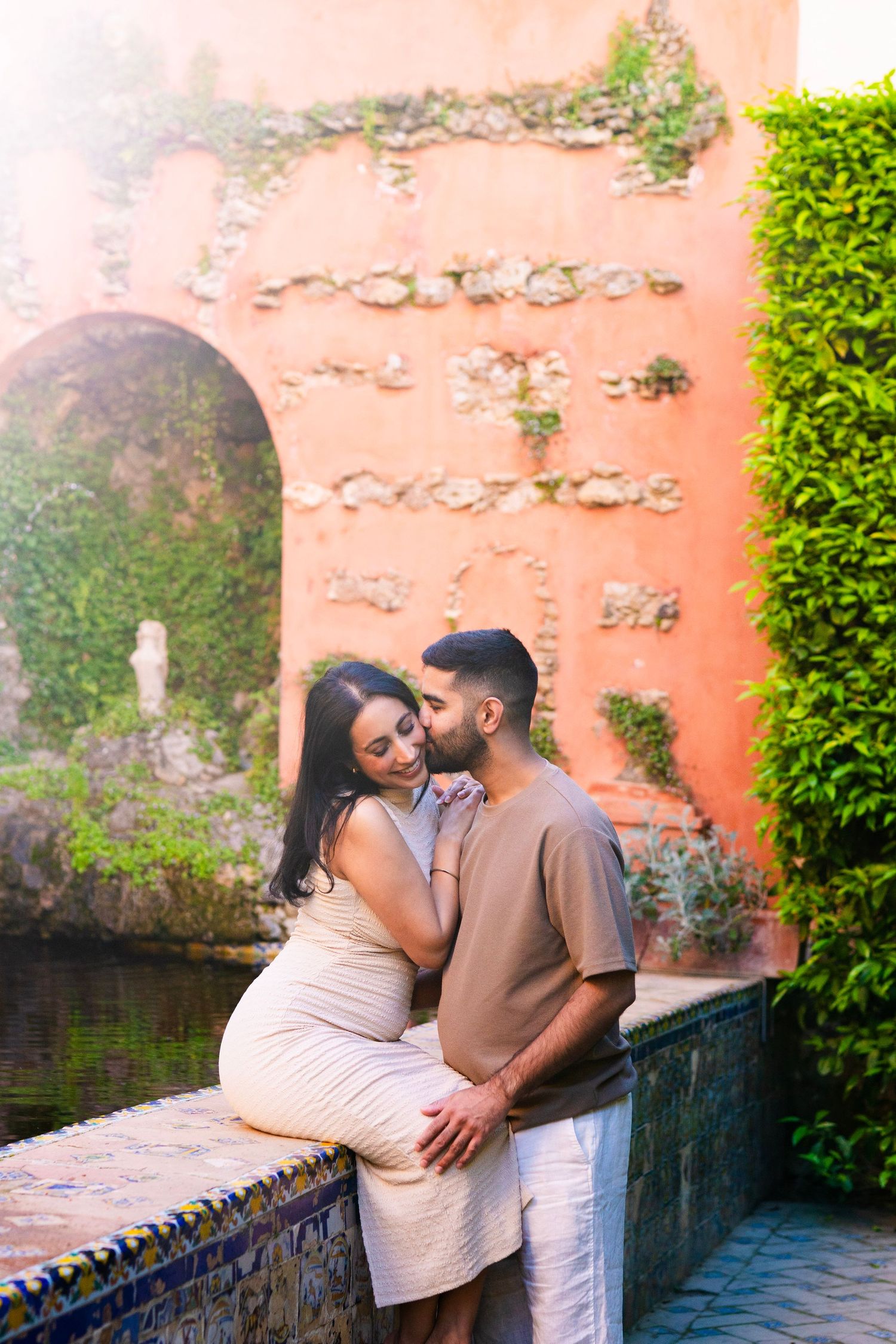 Romantic couple embracing by a tiled fountain with ivy-covered terracotta wall in a Mediterranean garden.
