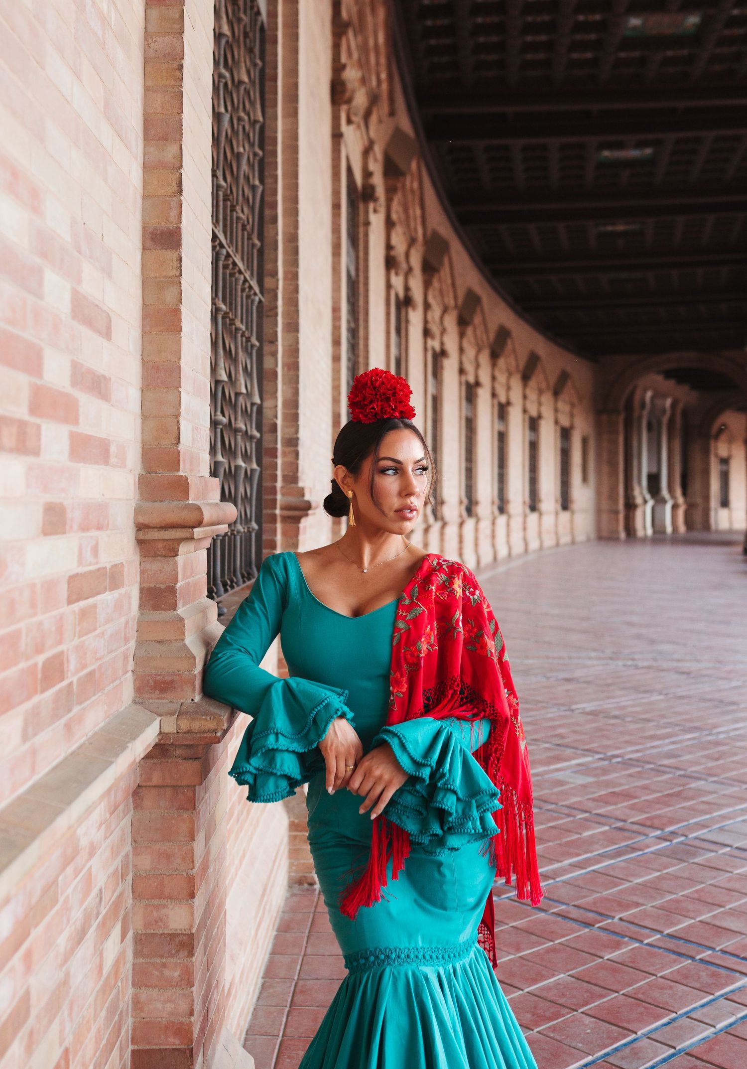 Flamenco dancer in teal ruffled dress with red shawl posing at Plaza de España arcade.