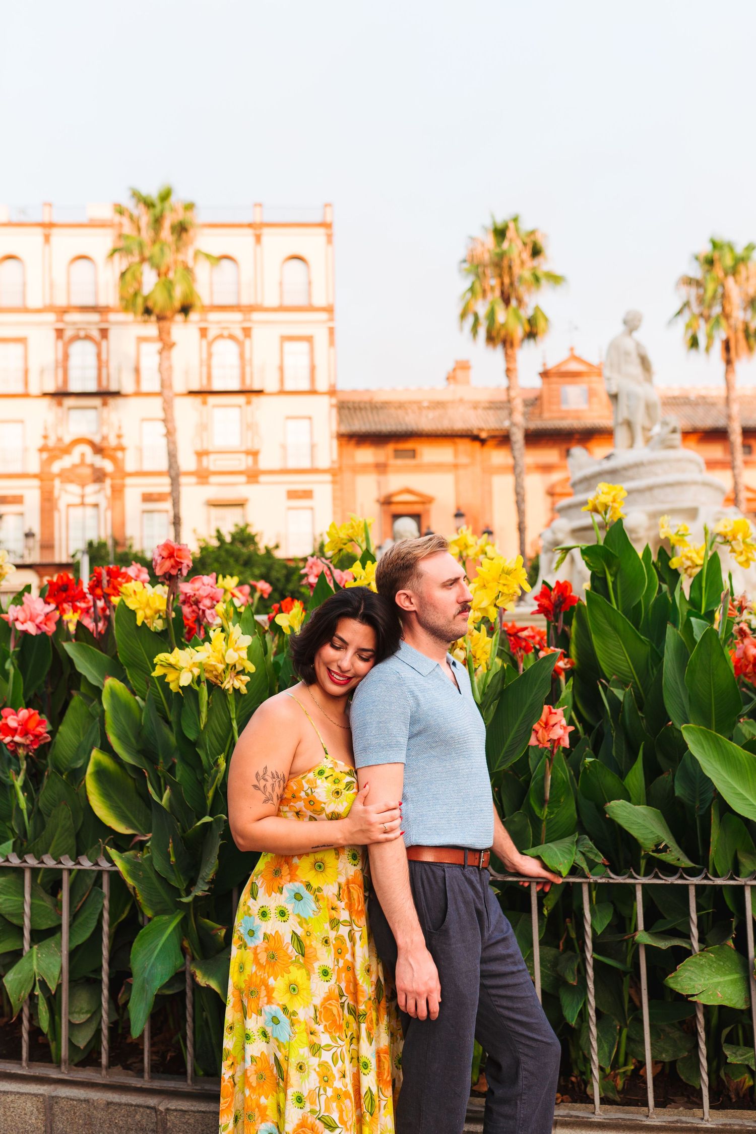 Couple embracing in front of colorful tropical flowers and a historic building with palm trees in warm golden light.