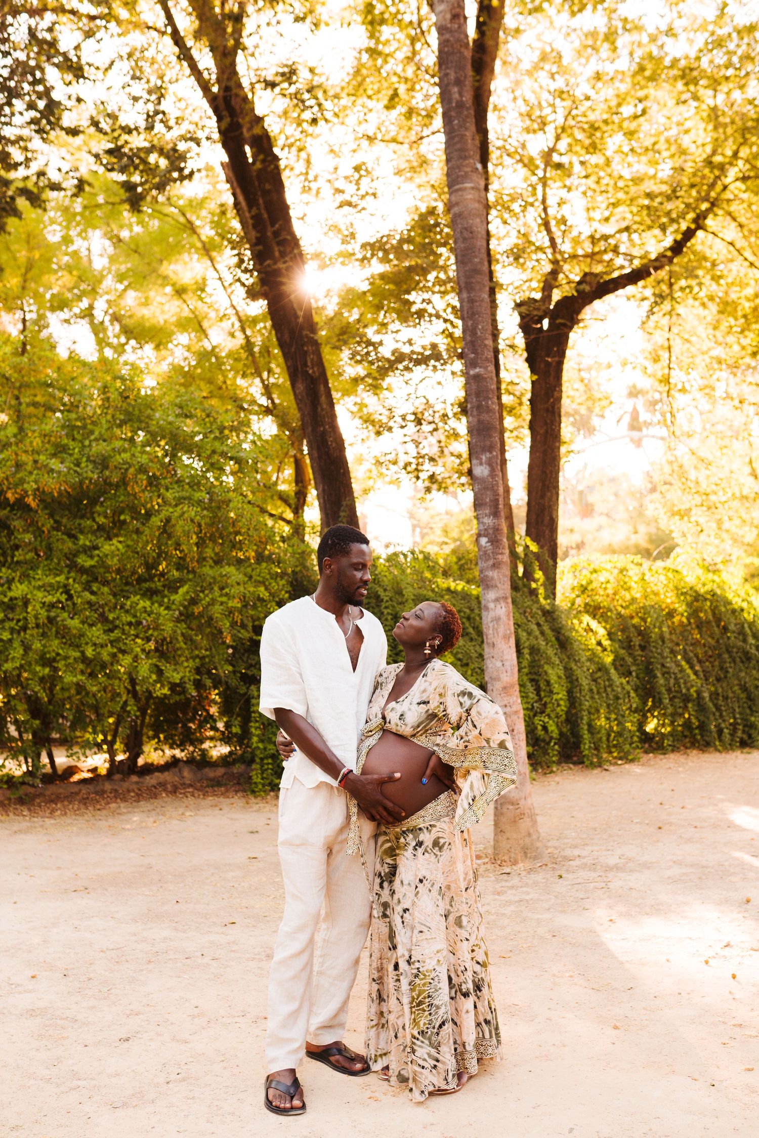 A pregnant couple shares a tender moment outdoors, bathed in golden sunlight among lush green trees.