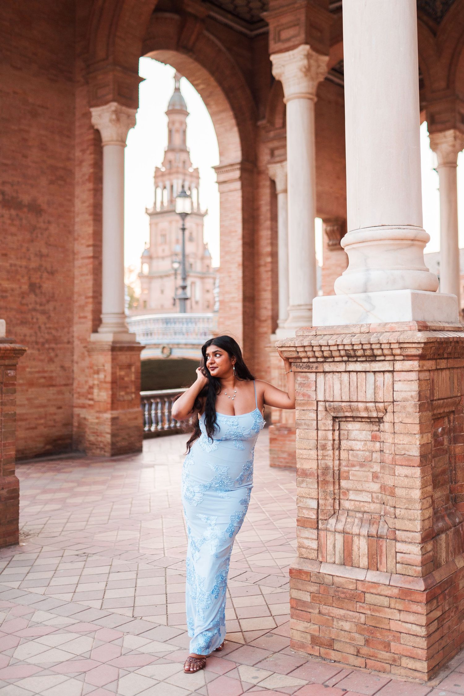 Woman in light blue maxi dress posing under brick arches at Plaza de España, Seville, Spain.