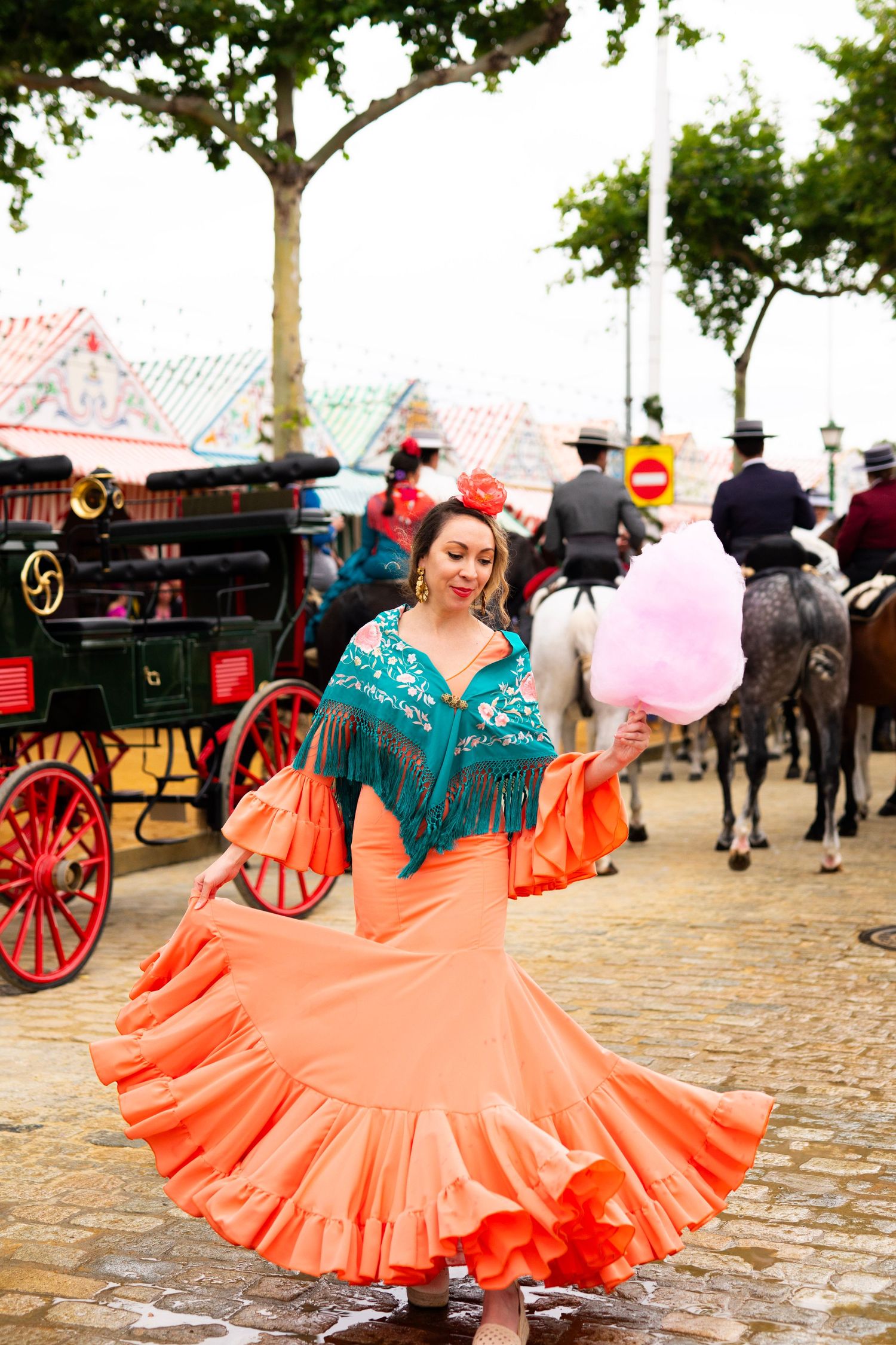 Woman in orange flamenco dress twirling with cotton candy near horse carriages at Spanish festival.