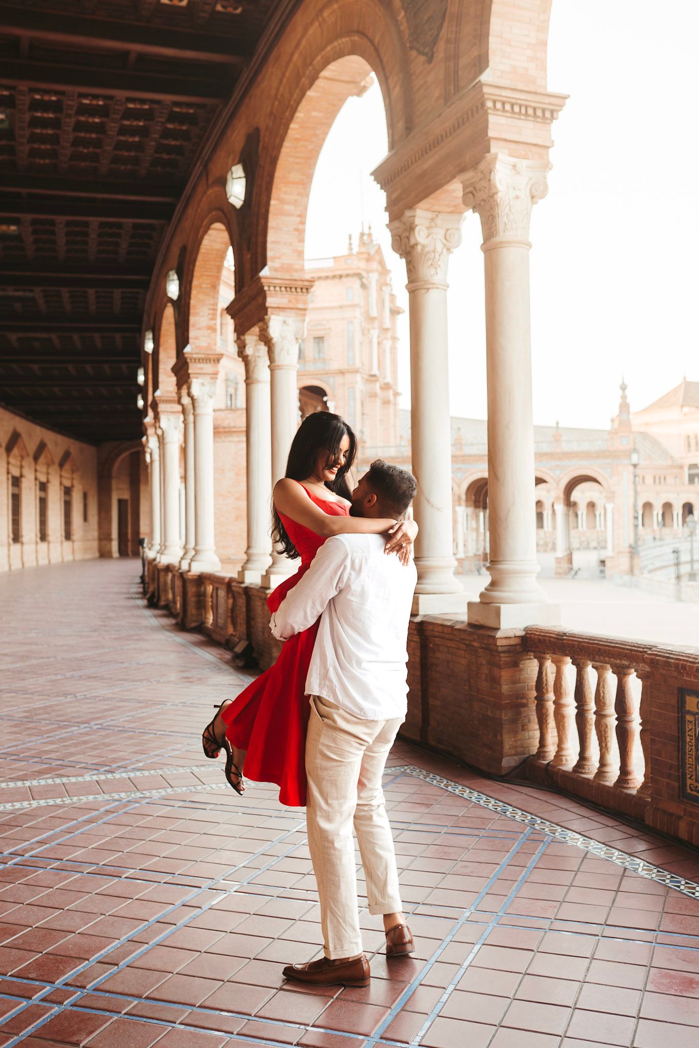 Man lifting woman in red dress under arched colonnade at Plaza de España, Seville, in a romantic embrace with sunlight streaming in.