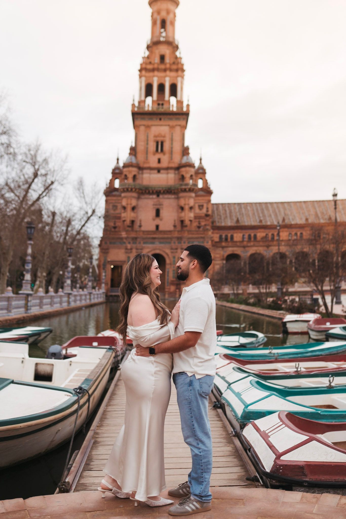 Romantic couple embracing on a dock with colorful boats at Plaza de España, Seville, Spain.