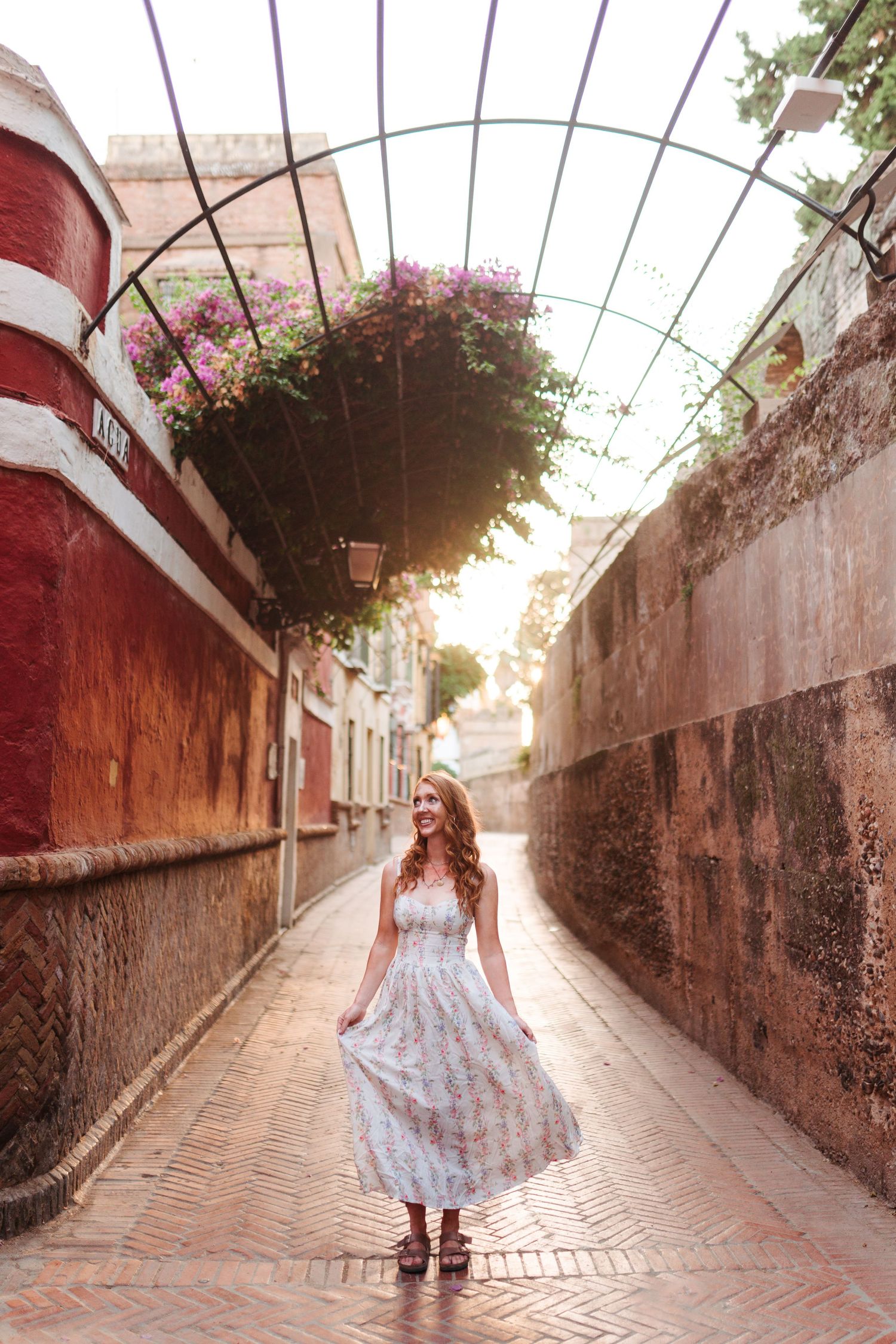 Woman in floral dress standing in a sunlit arched alleyway with colorful flowers and rustic stone walls.