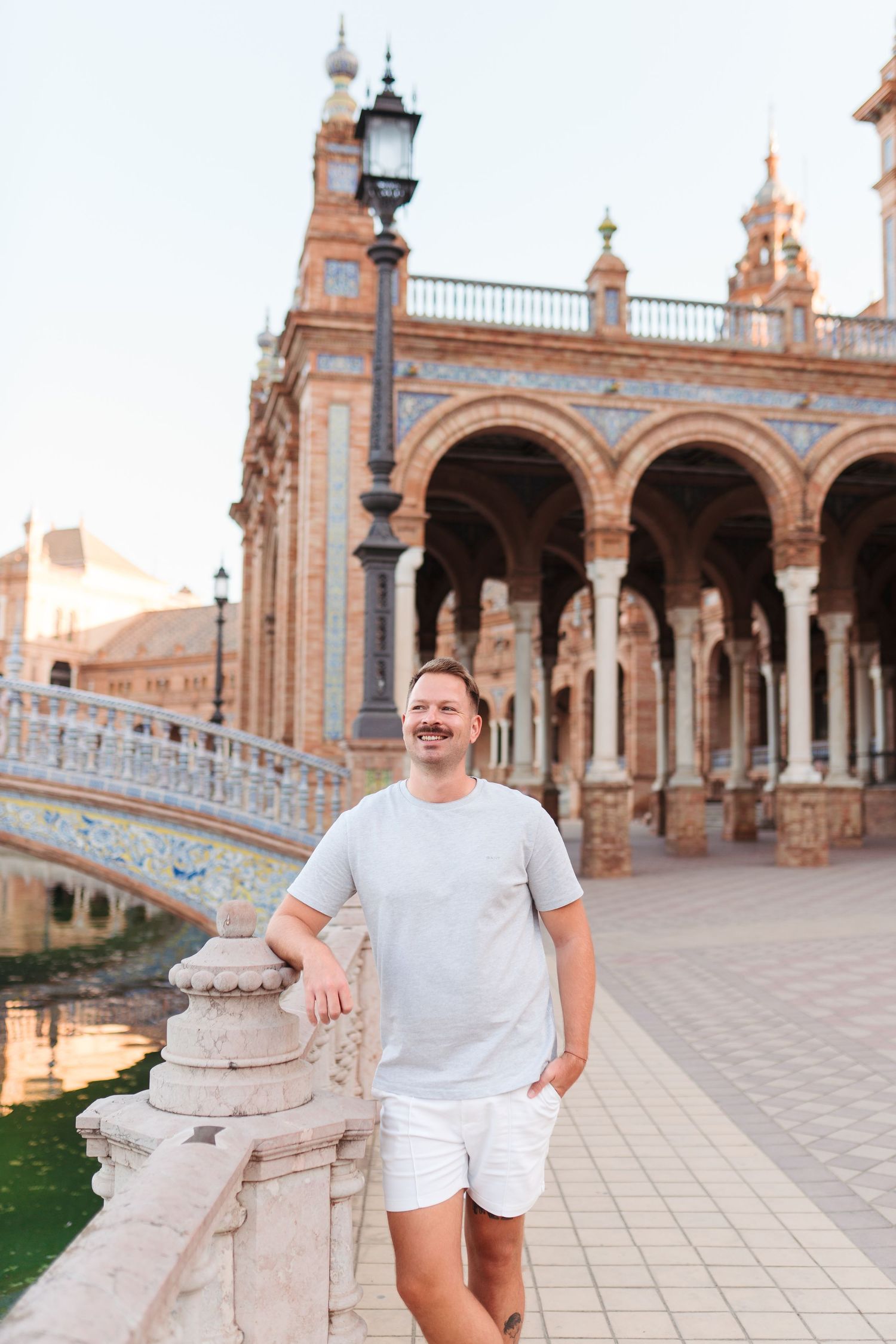 Smiling man in gray t-shirt and white shorts leaning on a stone railing at Plaza de España, Seville.