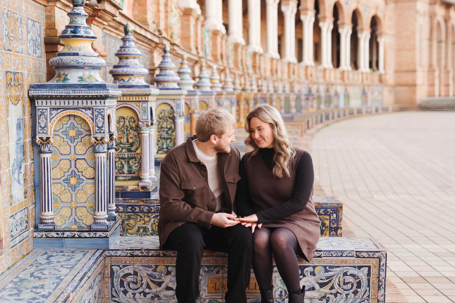 Engaged couple sitting together on tiled bench at Plaza de Espana admiring engagement ring.