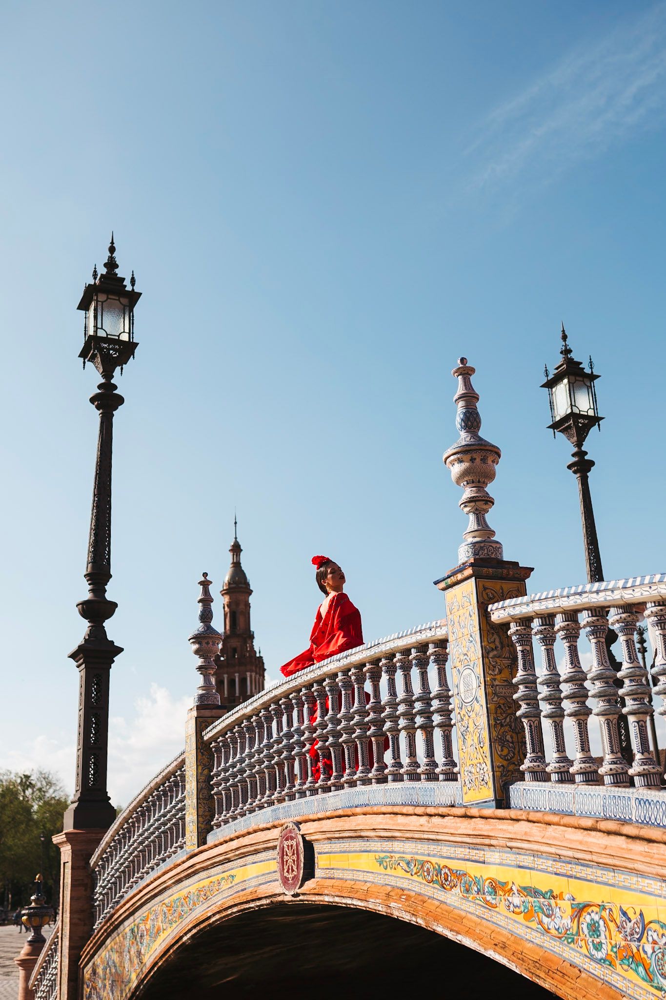 Flamenco dancer in red dress sitting on ornate tiled bridge at Plaza de España, Seville, Spain.