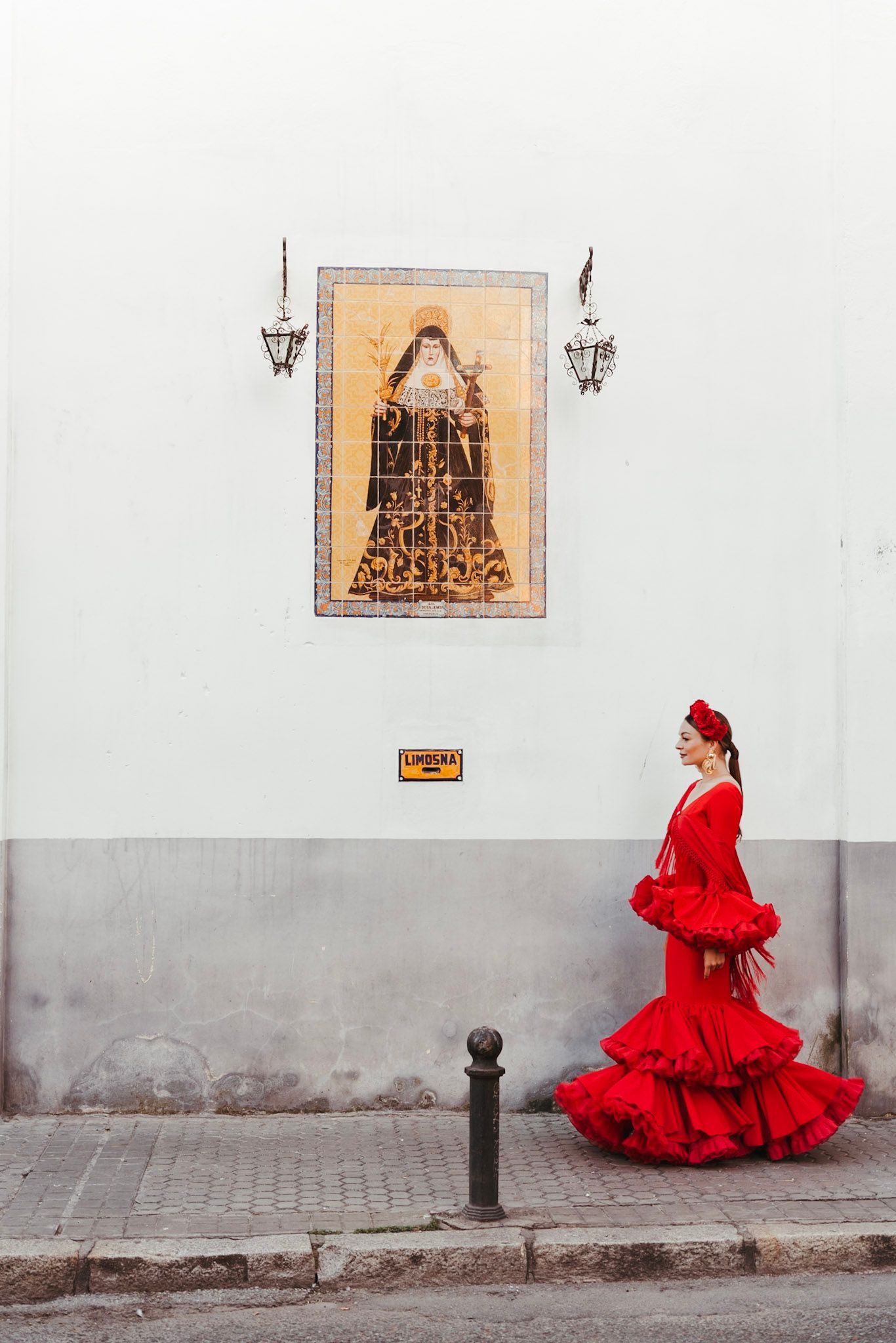 Woman in red flamenco dress walking past religious tile mural on white wall.
