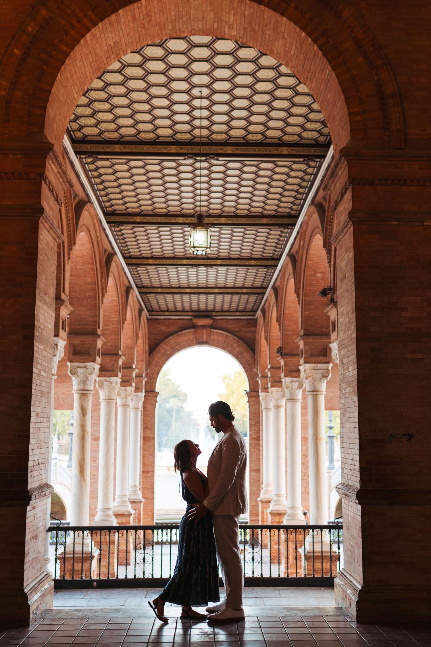 Couple silhouetted under ornate brick arch with honeycomb ceiling at Plaza de España, Seville, Spain.