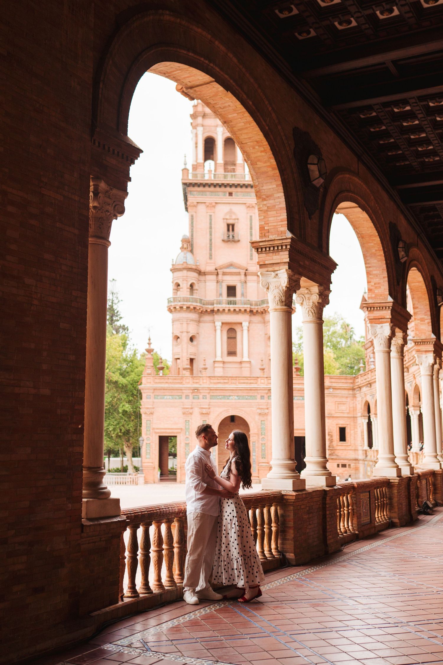 Couple embracing under ornate brick arches at Plaza de España, Seville, with a grand baroque tower in the background.