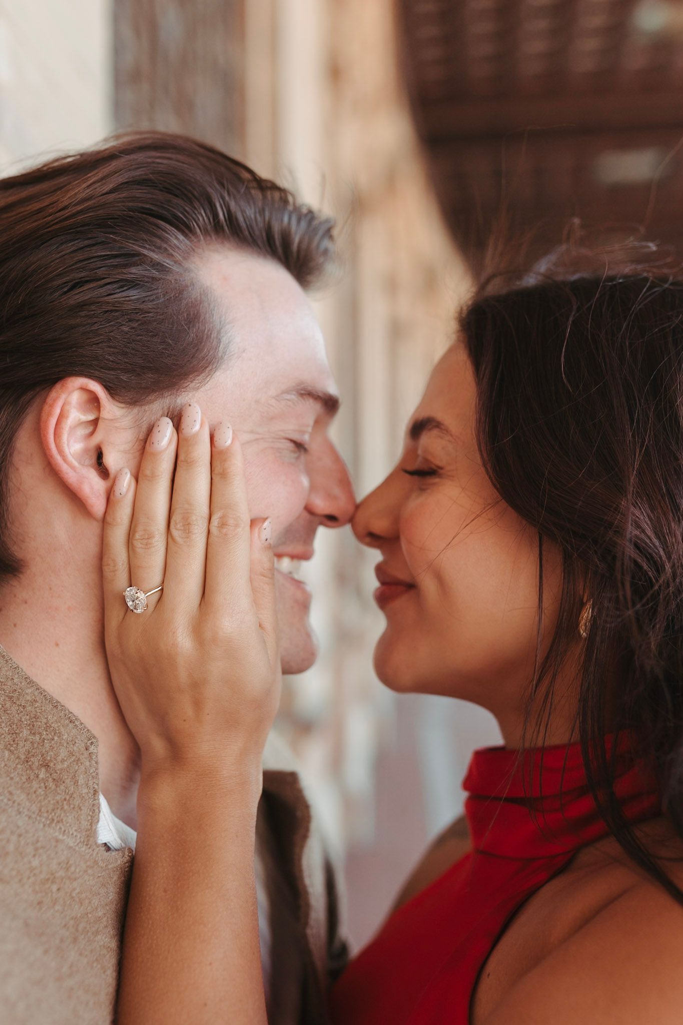 Close-up of smiling couple almost kissing, woman in red dress showing diamond engagement ring on hand.