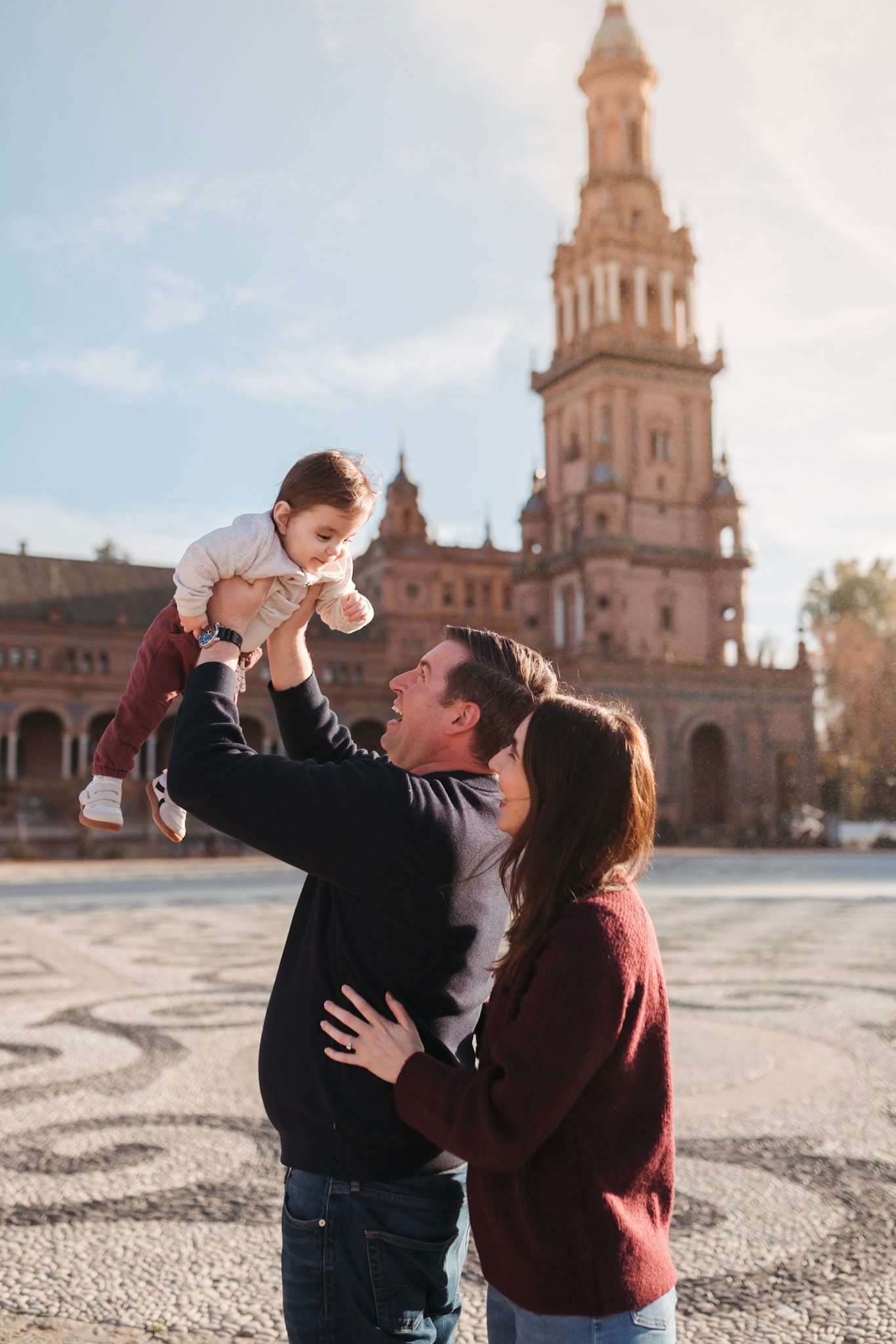 A happy couple lifts their baby in the air at Plaza de España, Seville, on a sunny day.