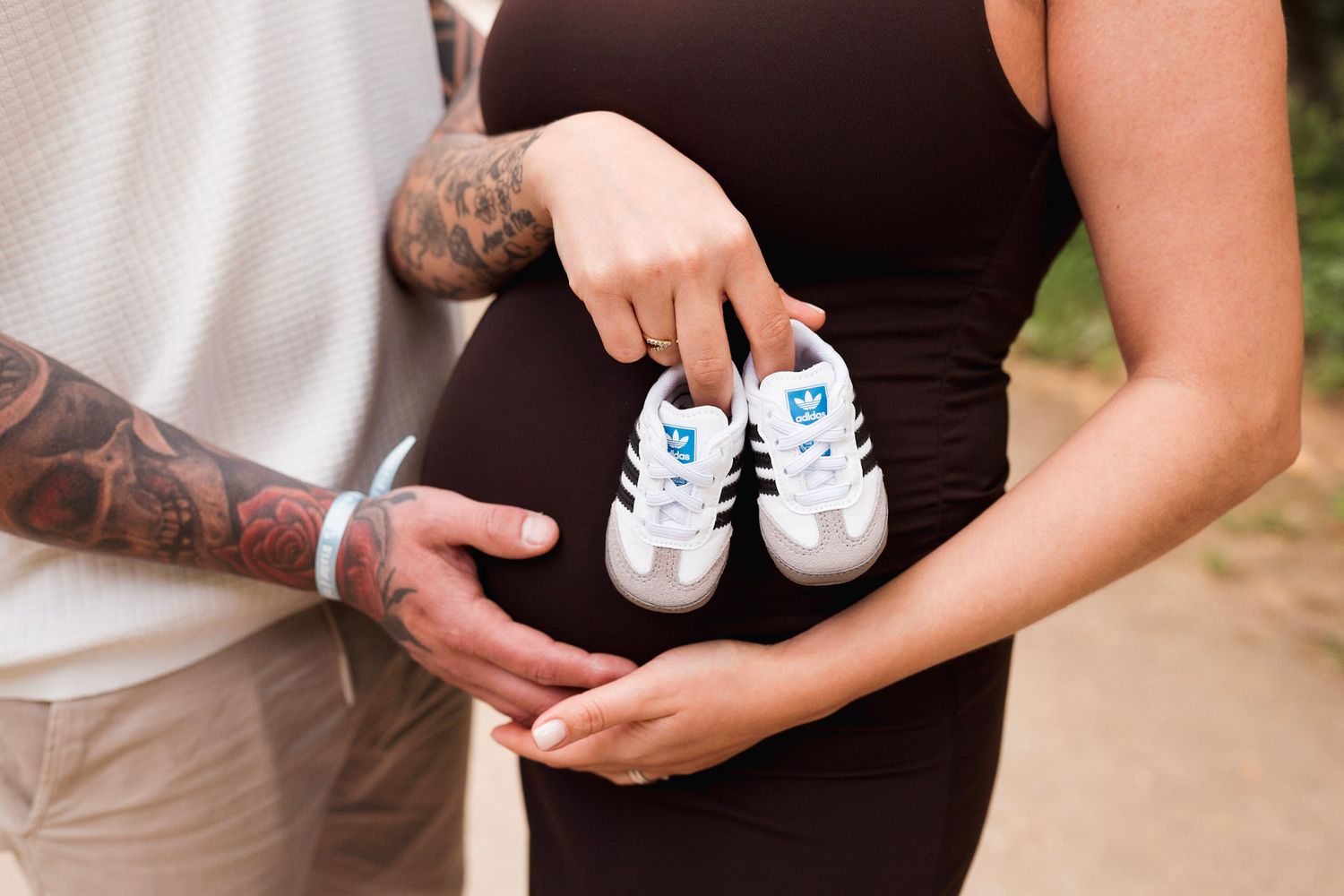 Couple holding tiny baby Adidas sneakers over pregnant belly, mans tattooed arm visible.