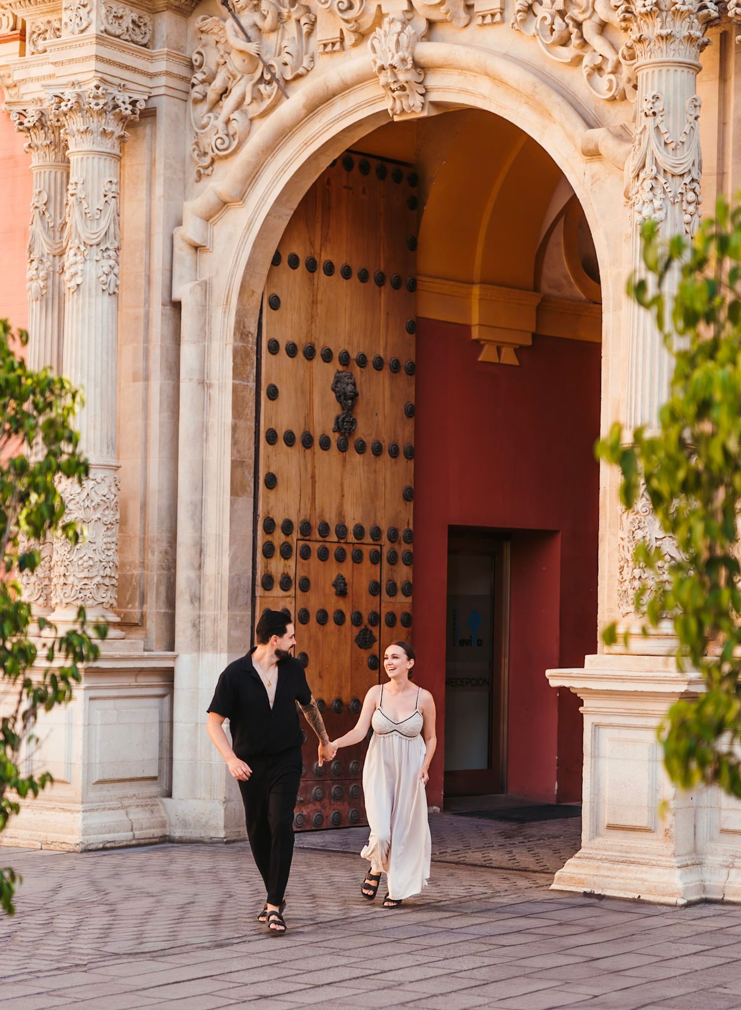 Couple holding hands walking under ornate stone arch doorway at Balboa Park San Diego.