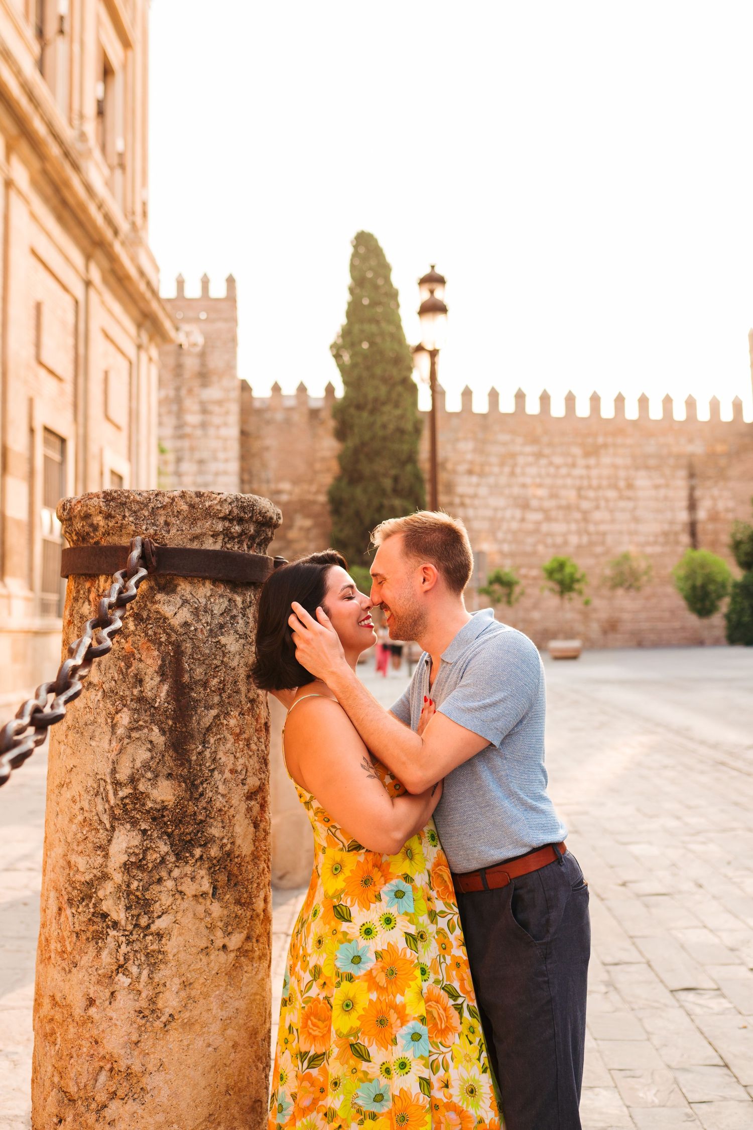 Couple sharing a romantic moment by a stone pillar in a sunlit historic European plaza.