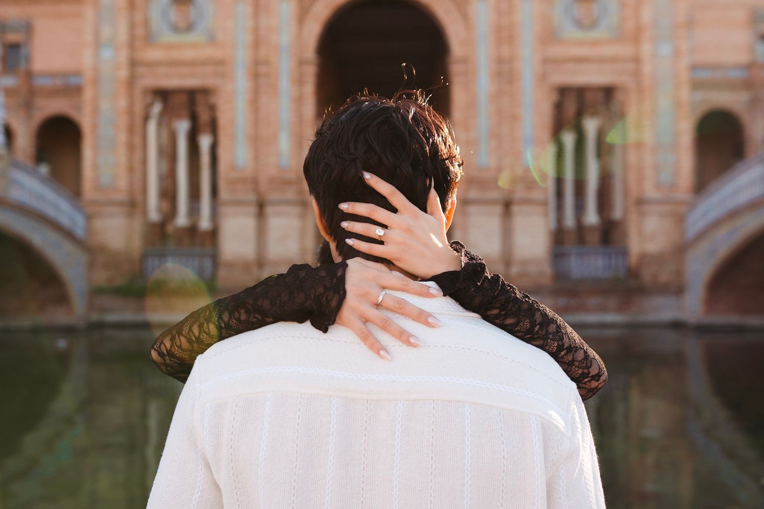 Couple embracing from behind at a historic architectural landmark, woman wearing diamond engagement ring.
