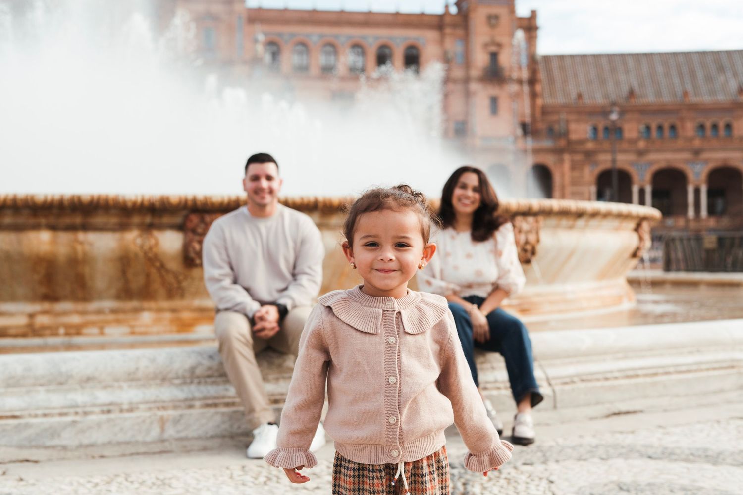 Adorable toddler smiles in front of the Plaza de España fountain with parents sitting behind her.