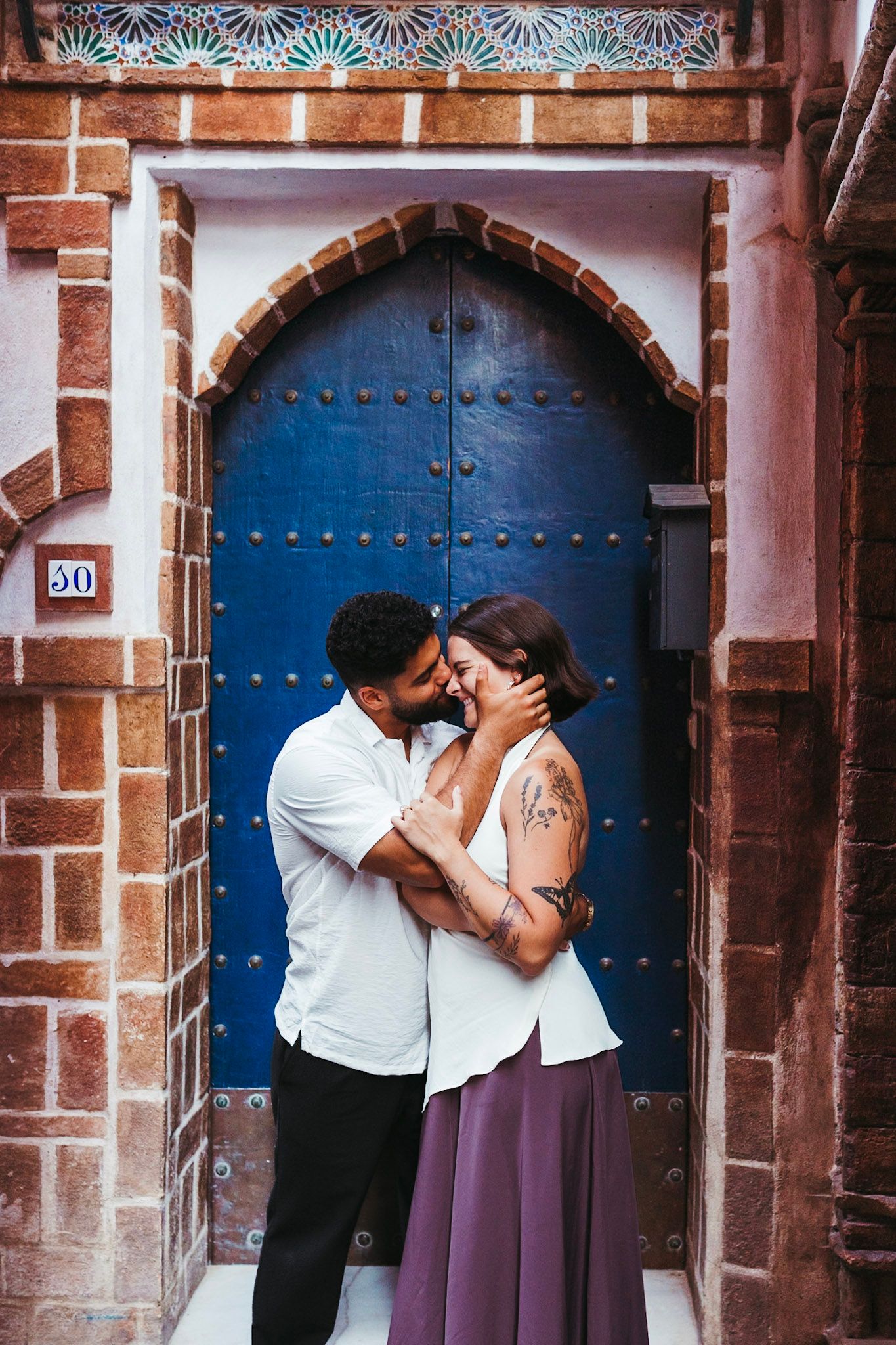 Couple sharing intimate kiss in front of ornate blue wooden door with Moorish brick archway.