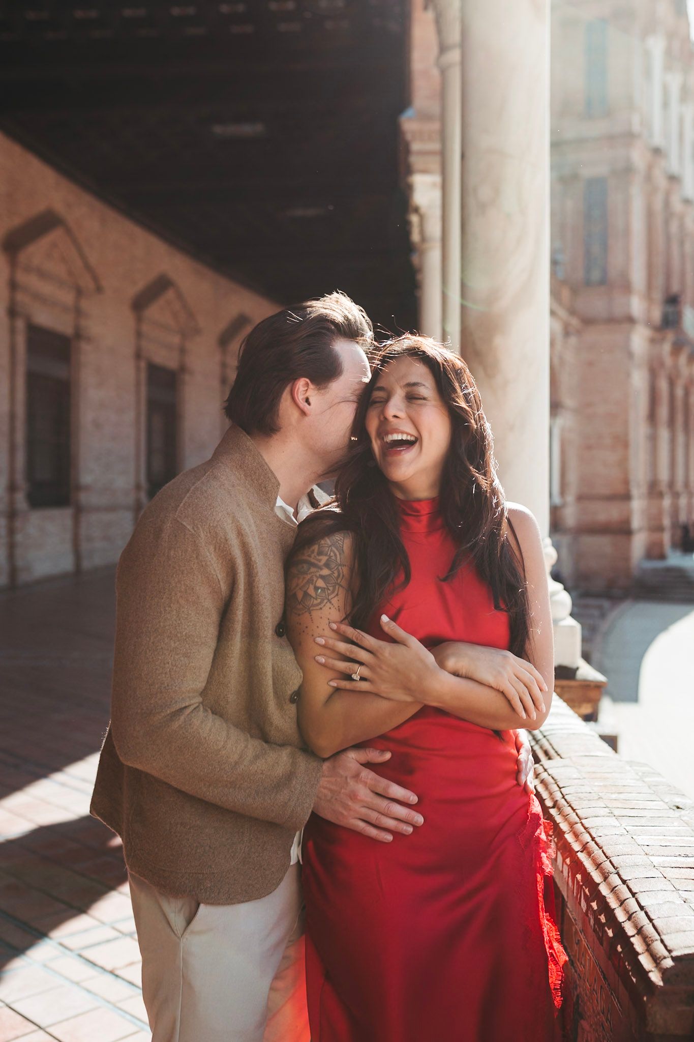 Man whispering to laughing woman in red dress at Plaza de España colonnade, Seville.