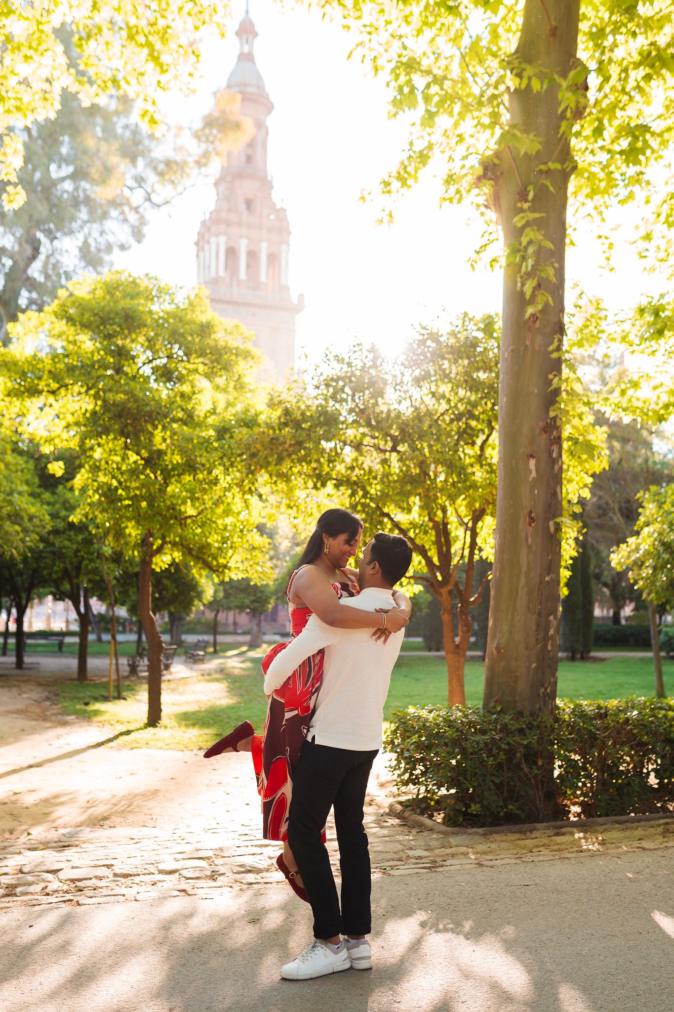 Man lifts woman in red dress among sunlit trees near Plaza de Espana tower in Seville, Spain.