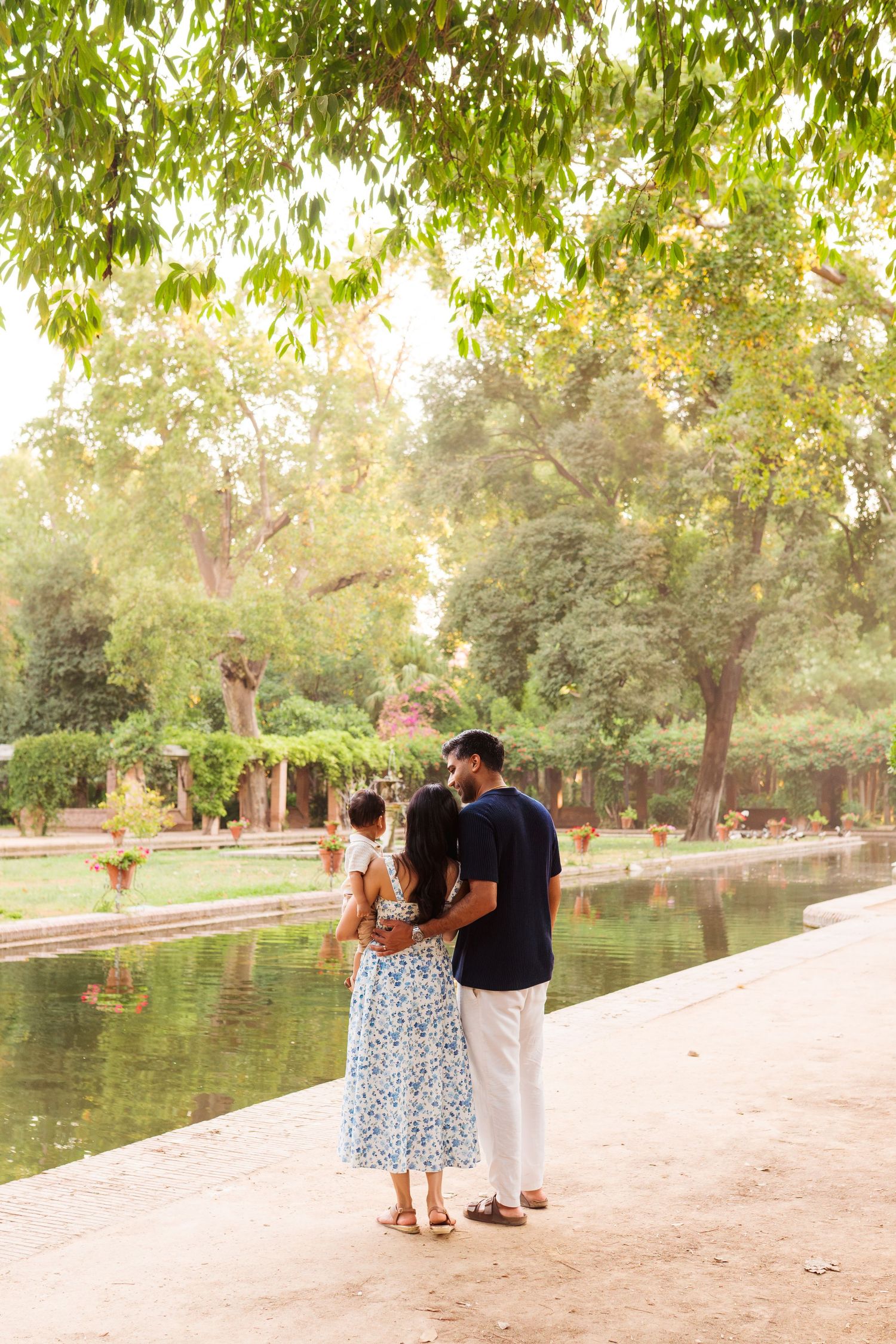 A family of three stands by a serene pond in a lush green park, with the couple holding their young child.