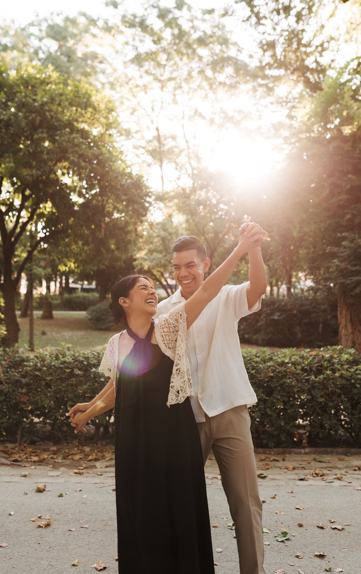 Happy couple dancing outdoors in a sunlit park, laughing and holding hands raised, surrounded by lush green trees.