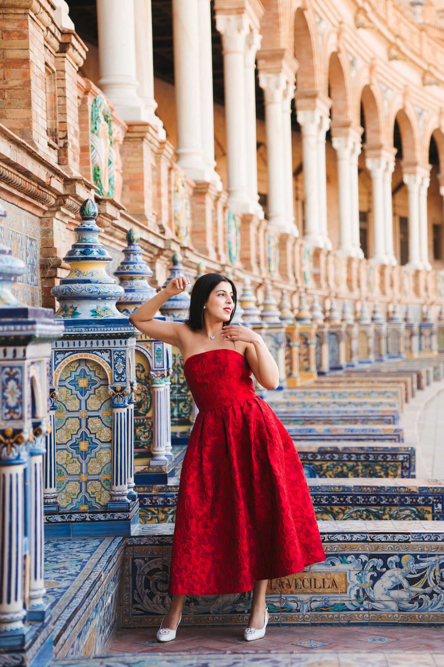 Woman in red strapless gown poses at Plaza de España Seville, surrounded by ornate tilework and arched colonnade.