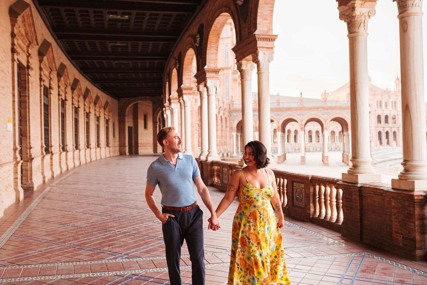 Couple holding hands walking through ornate arched colonnade at Plaza de España, Seville, Spain.