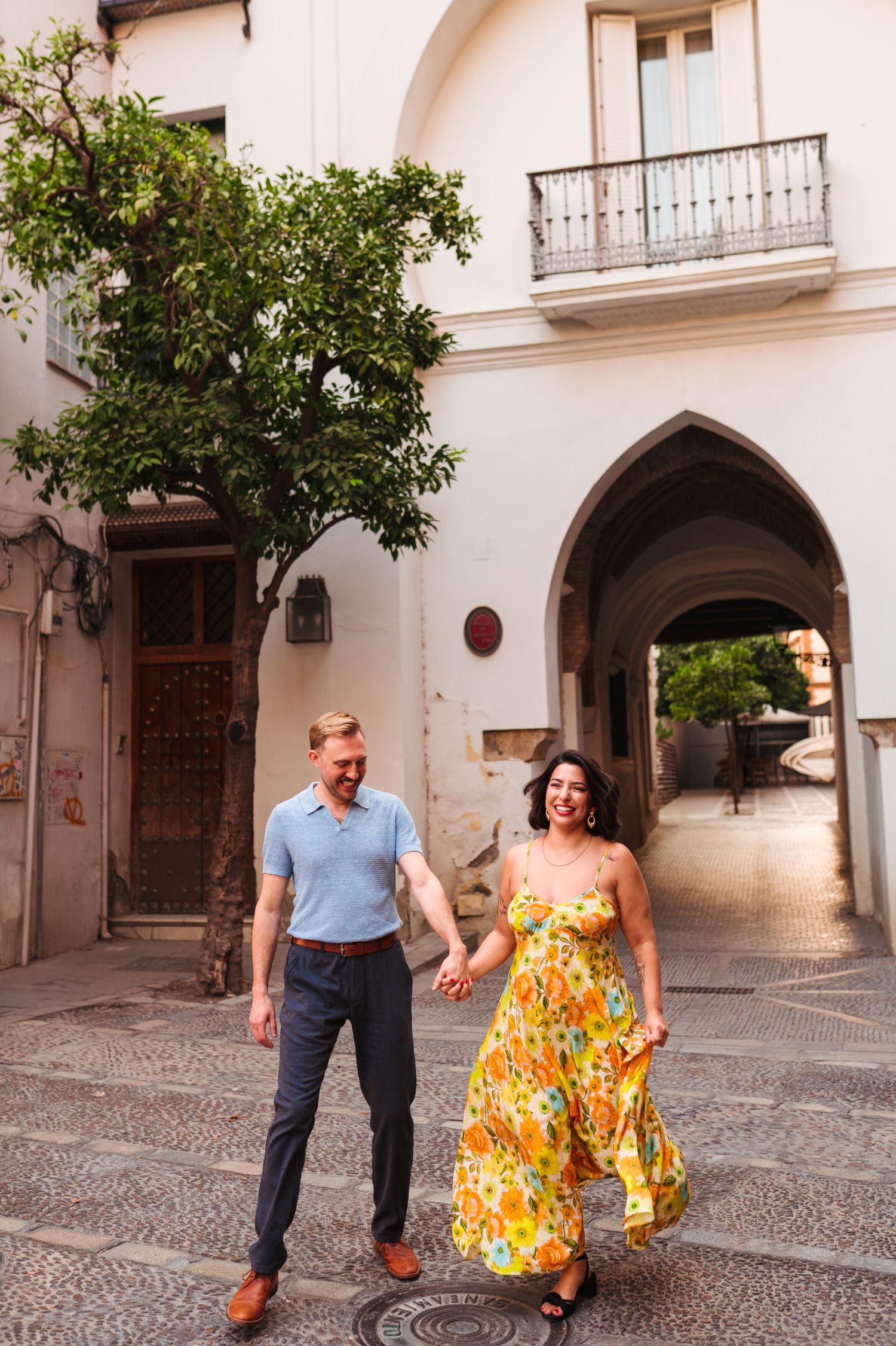 Couple holding hands and walking through a charming European cobblestone alley, woman in floral yellow dress.