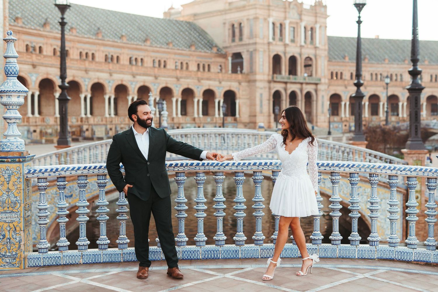 Couple holding hands on a tiled bridge at Plaza de España, Seville, with grand baroque architecture behind.