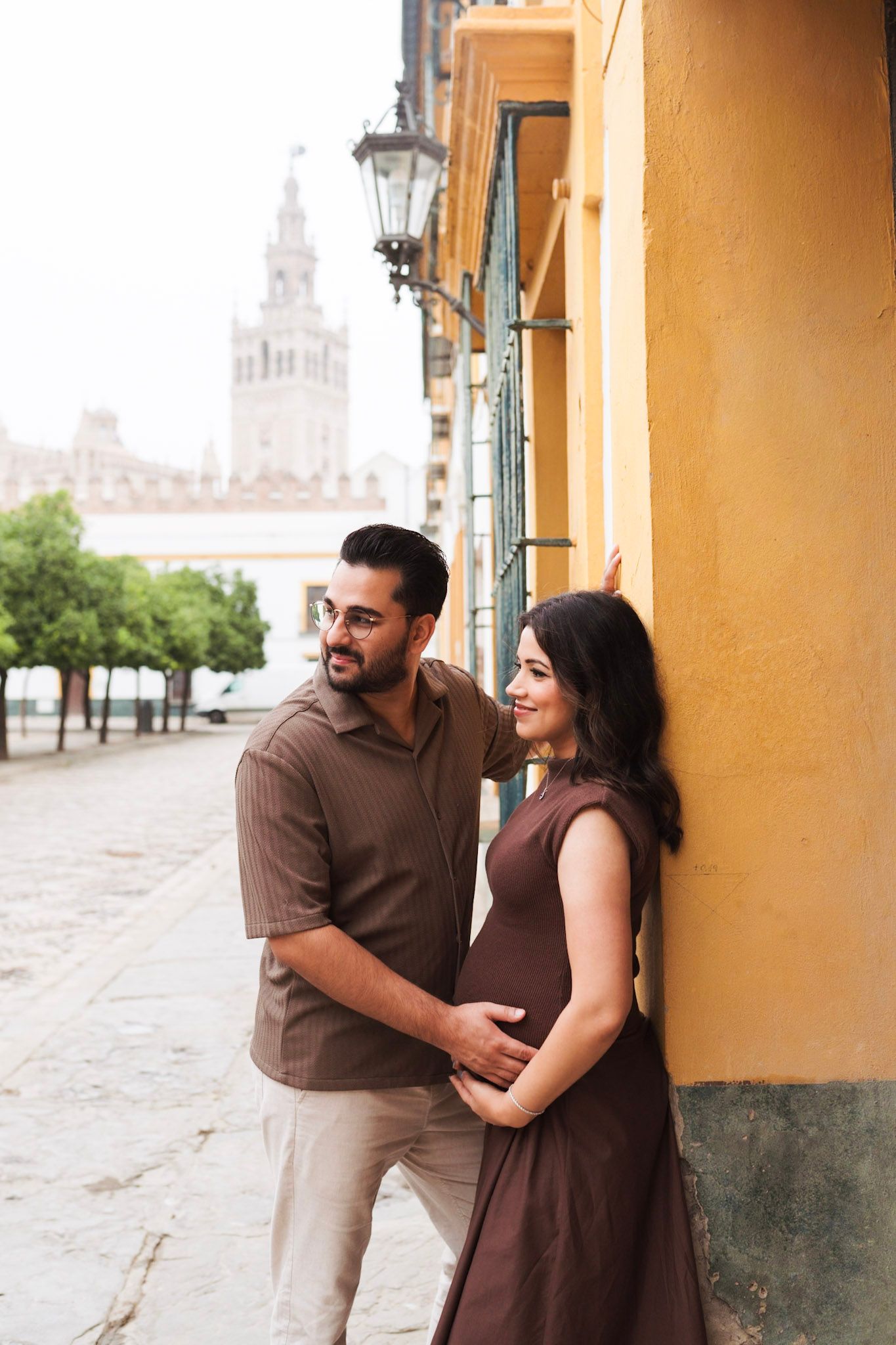 Pregnant couple embracing against yellow wall with Giralda tower in background, Seville.
