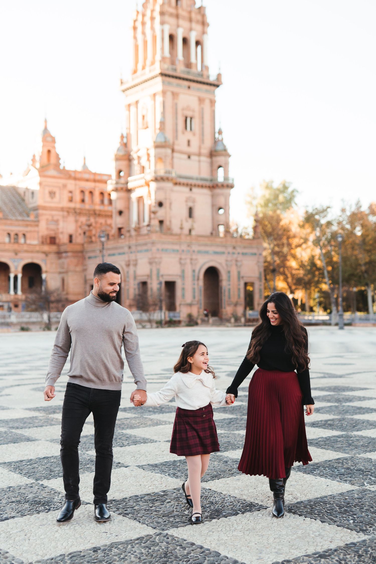 Happy family of three holds hands while walking at Plaza de España in Seville at sunset.