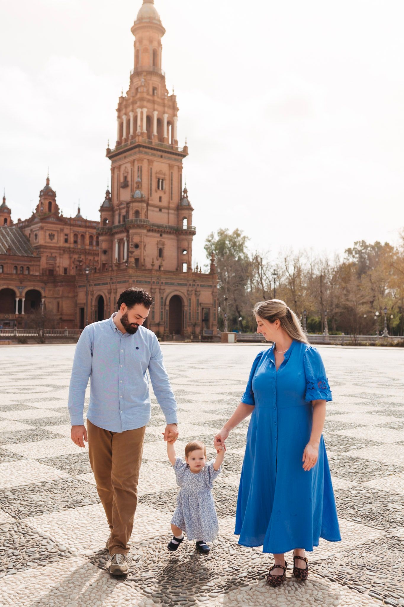 A family of three walks hand-in-hand across the mosaic plaza at Plaza de España, Seville, Spain.