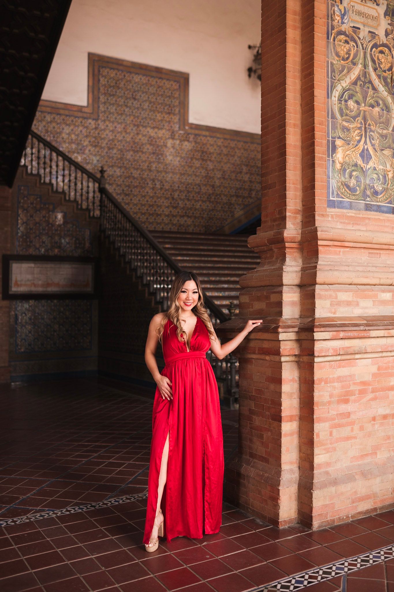 Woman in red dress stands by ornate tiled column with grand staircase in Seville.