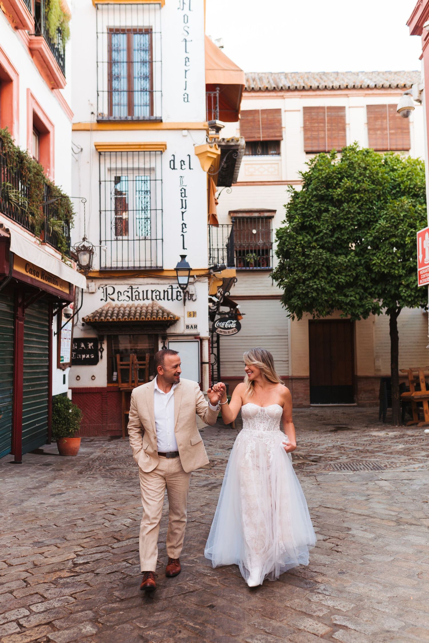 Bride and groom walk hand-in-hand through a charming cobblestone plaza in Seville, Spain, near a historic restaurant.