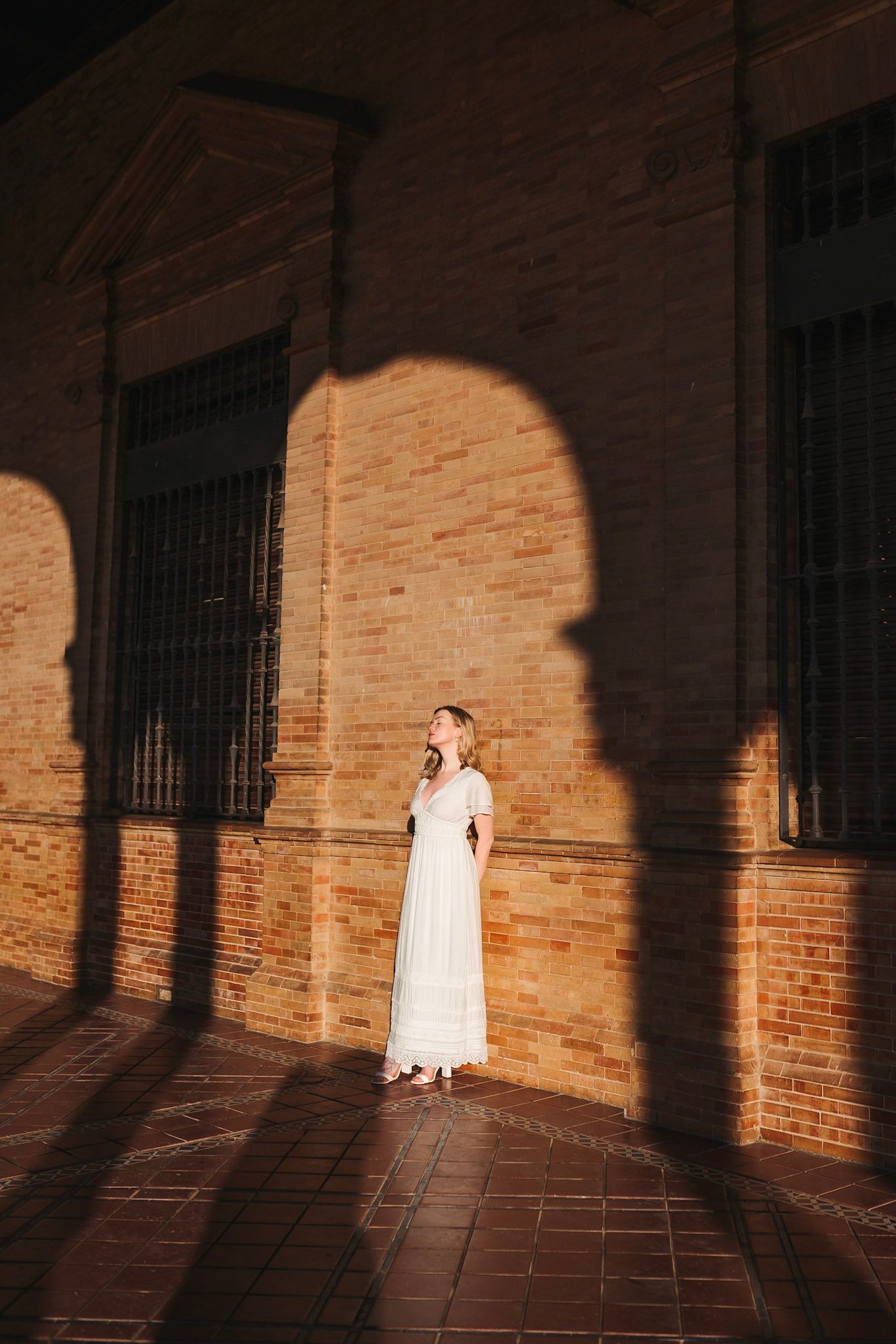 Woman in white dress stands in dramatic arched shadow on brick wall.