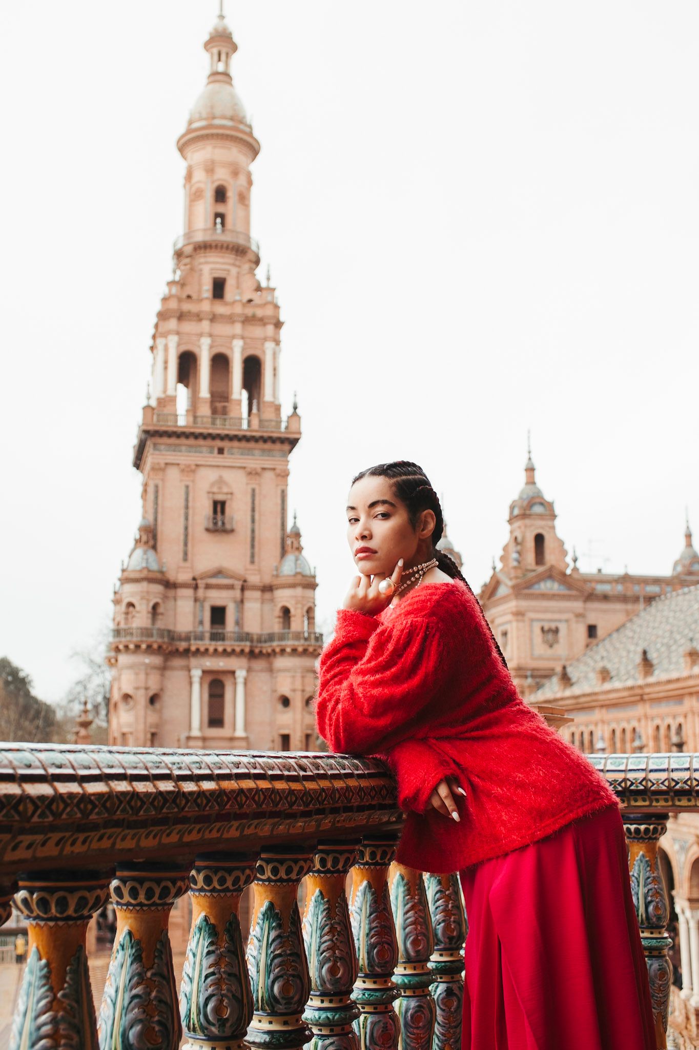 Young woman in red outfit leans on ornate railing at Plaza de España, Seville, with iconic tower behind.