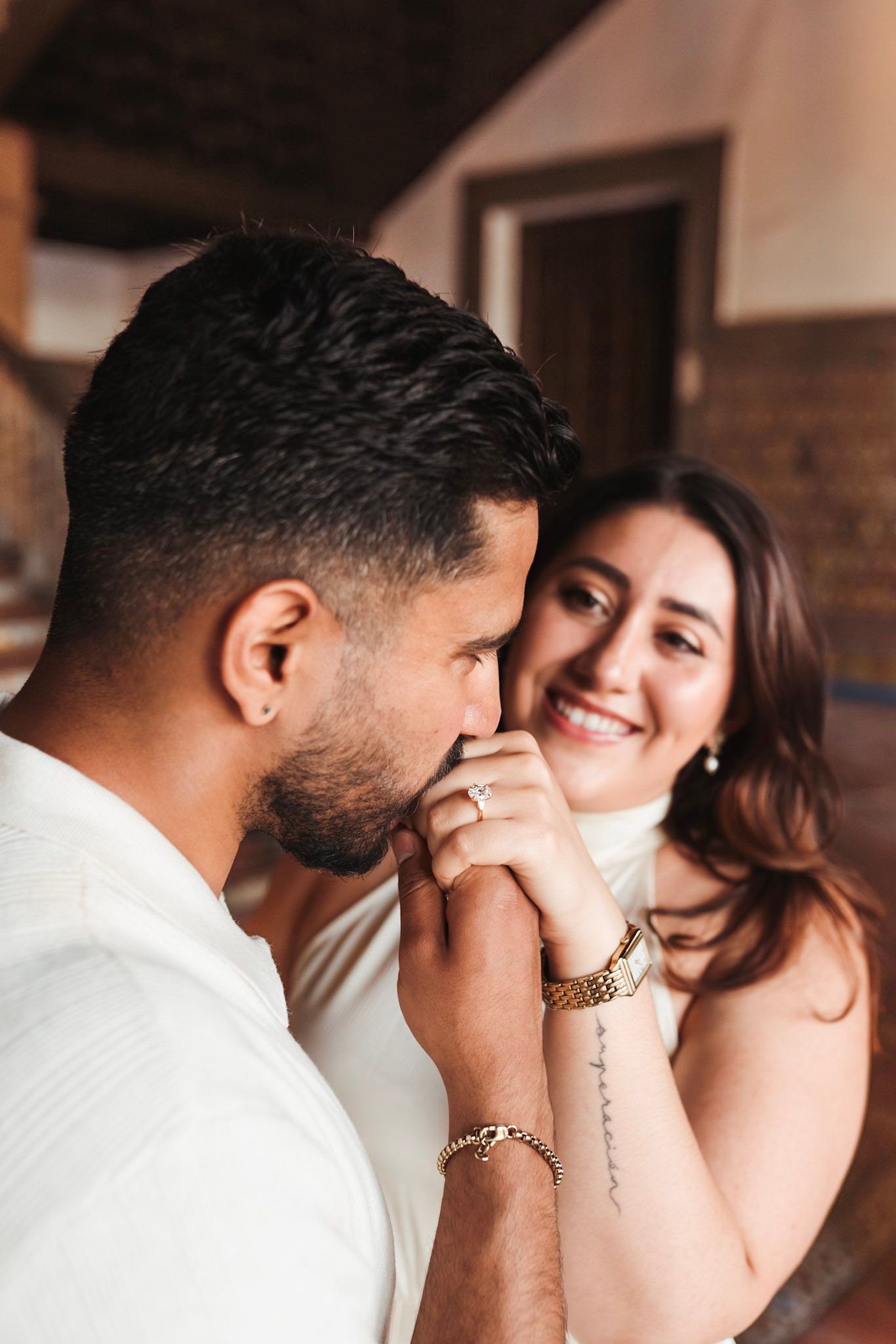 Man kisses fiancée's hand with engagement ring as she smiles warmly in rustic Seville interior.