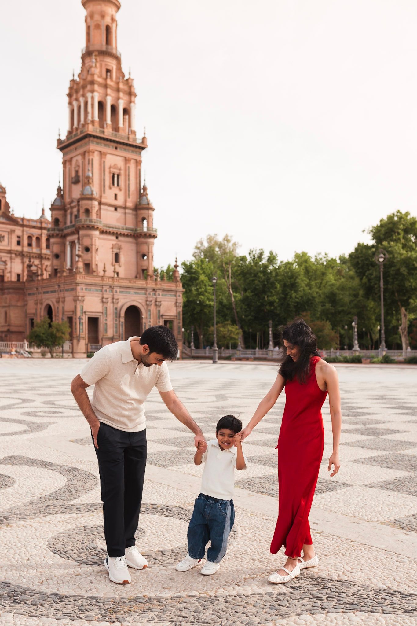 Parents in red dress and white shirt hold hands with their young son at Plaza de Espana Seville.