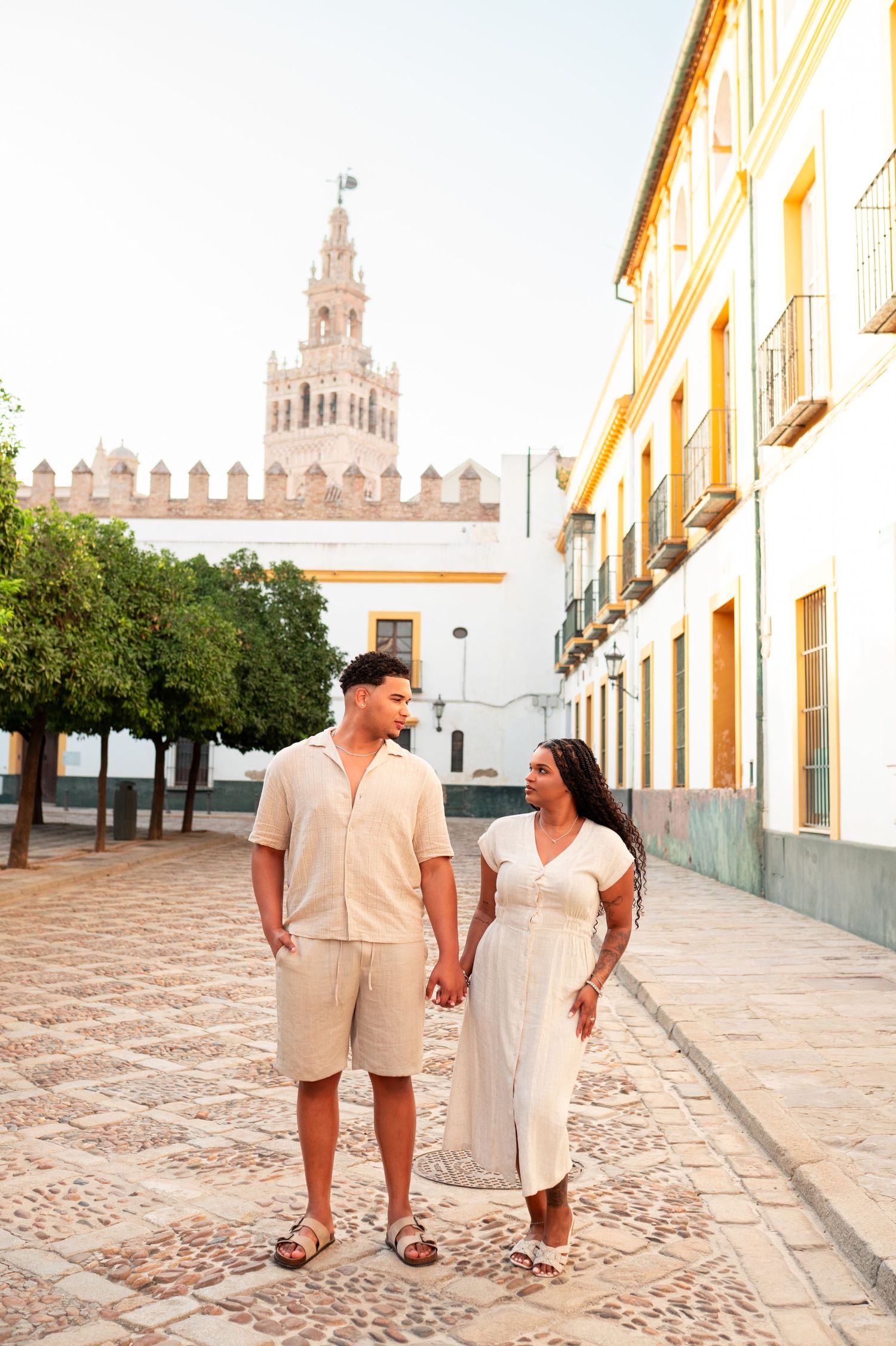 Couple holding hands in a sunny cobblestone plaza with the Giralda tower of Seville, Spain in the background.