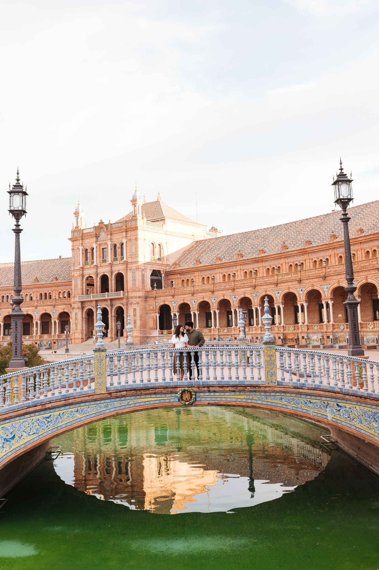 Couple standing on ornate tiled bridge at Plaza de España, Seville, with green canal and grand arcade reflected in water.