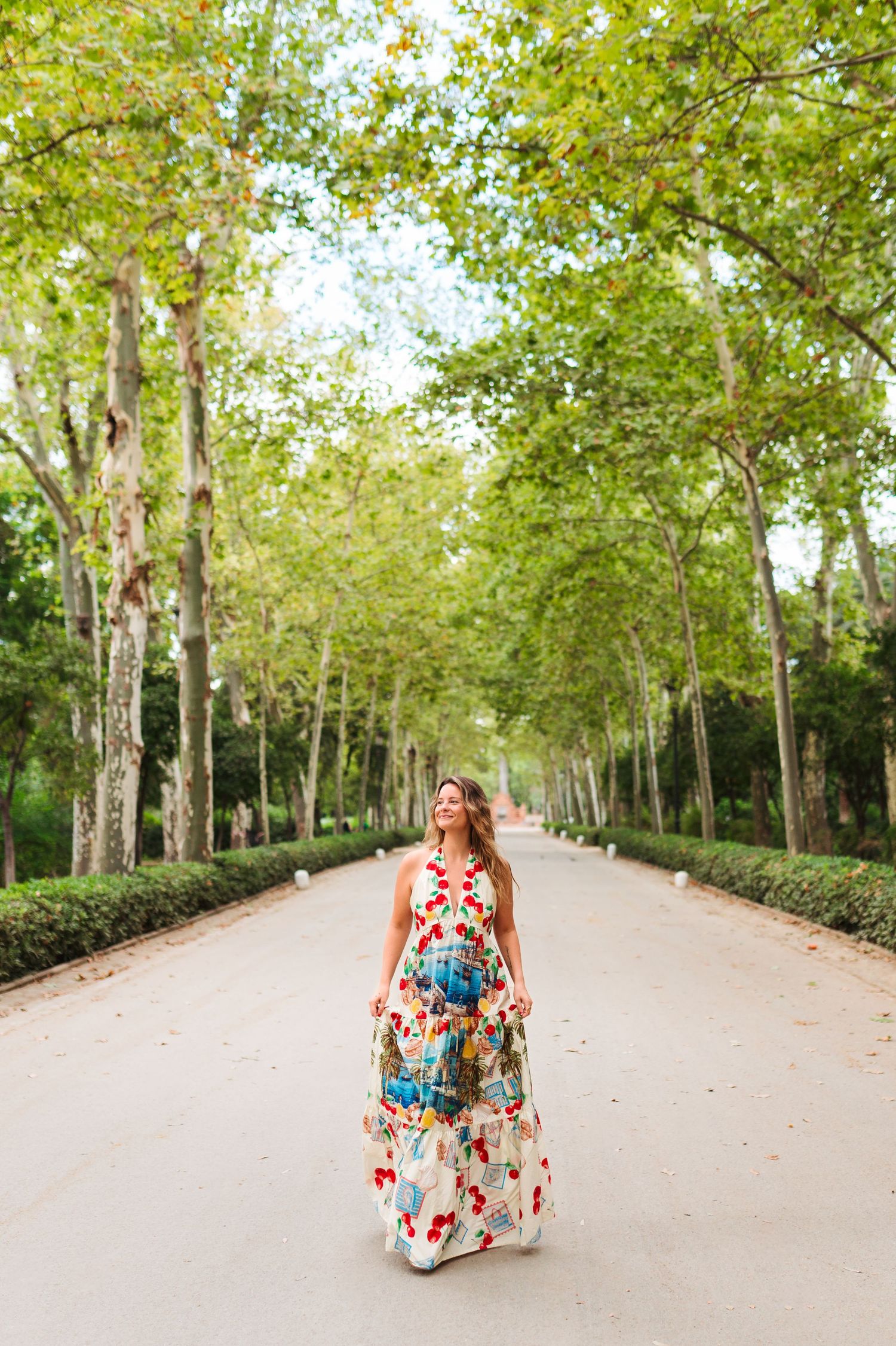 Woman in floral maxi dress walking along tree-lined park path, looking up at lush green canopy overhead.